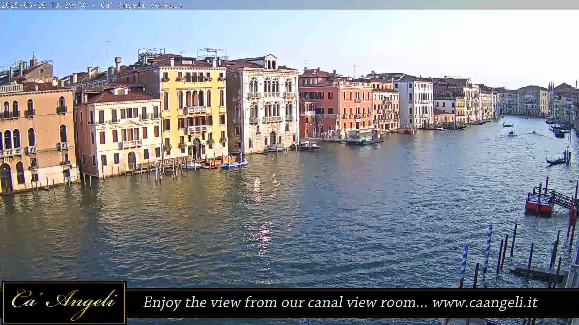 A sunny view of a wide canal in Venice, Italy, lined with tall, colorful buildings, with several boats on the water.