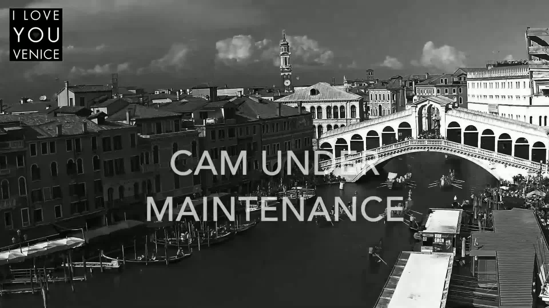 A black and white, high-angle view shows a canal in Venice, Italy, with a bridge, boats, and buildings under a partly cloudy sky.