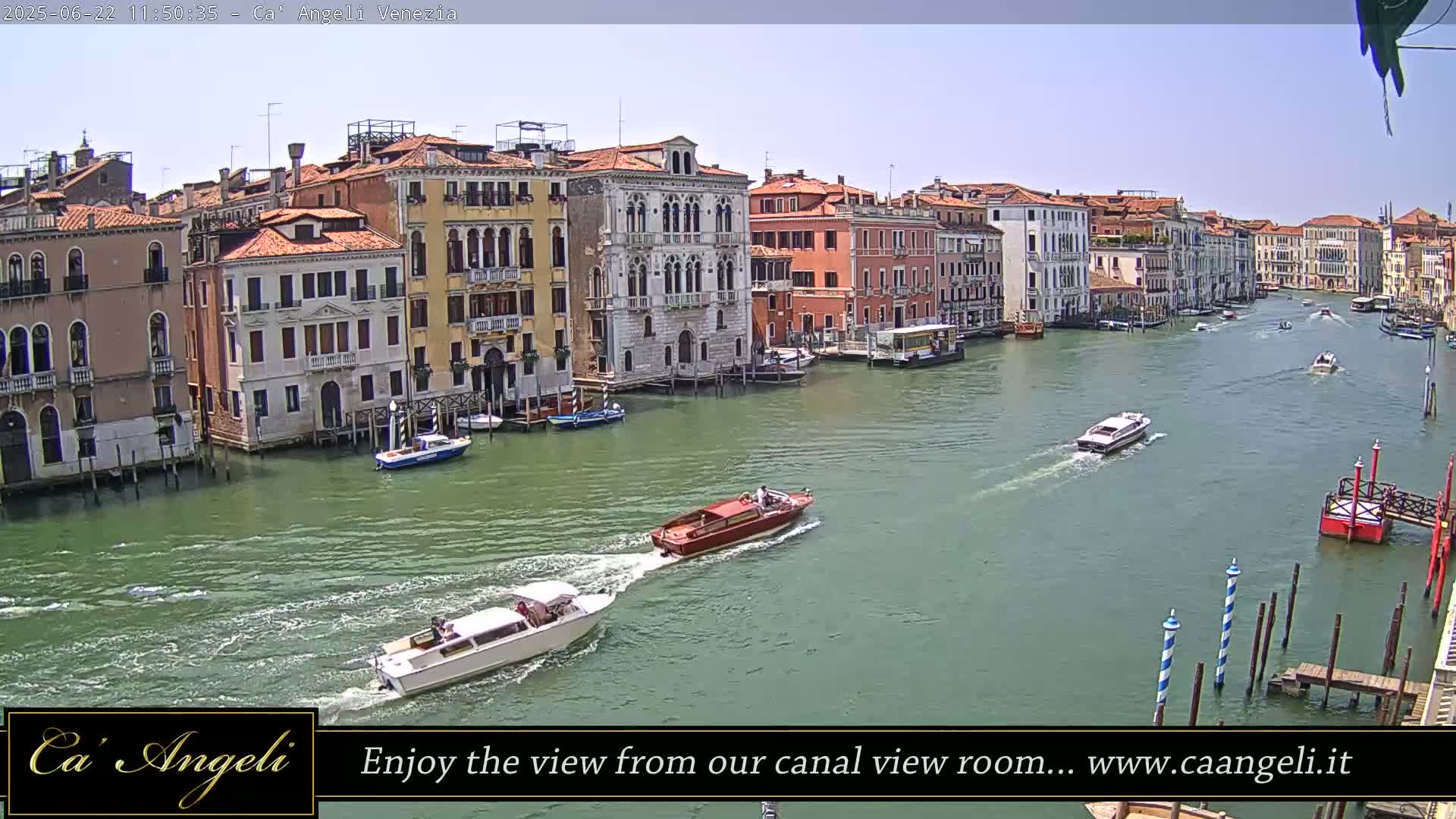 Several boats travel on a wide canal lined with tall, colorful buildings under a mostly sunny sky.