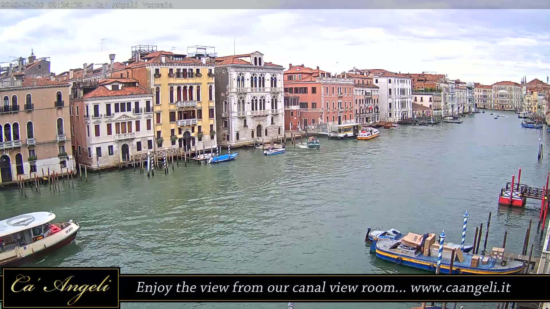 A wide shot of a canal in Venice, Italy, shows several multi-story buildings lining the waterway under a cloudy sky, with various boats moored along the buildings and others traveling on the water.