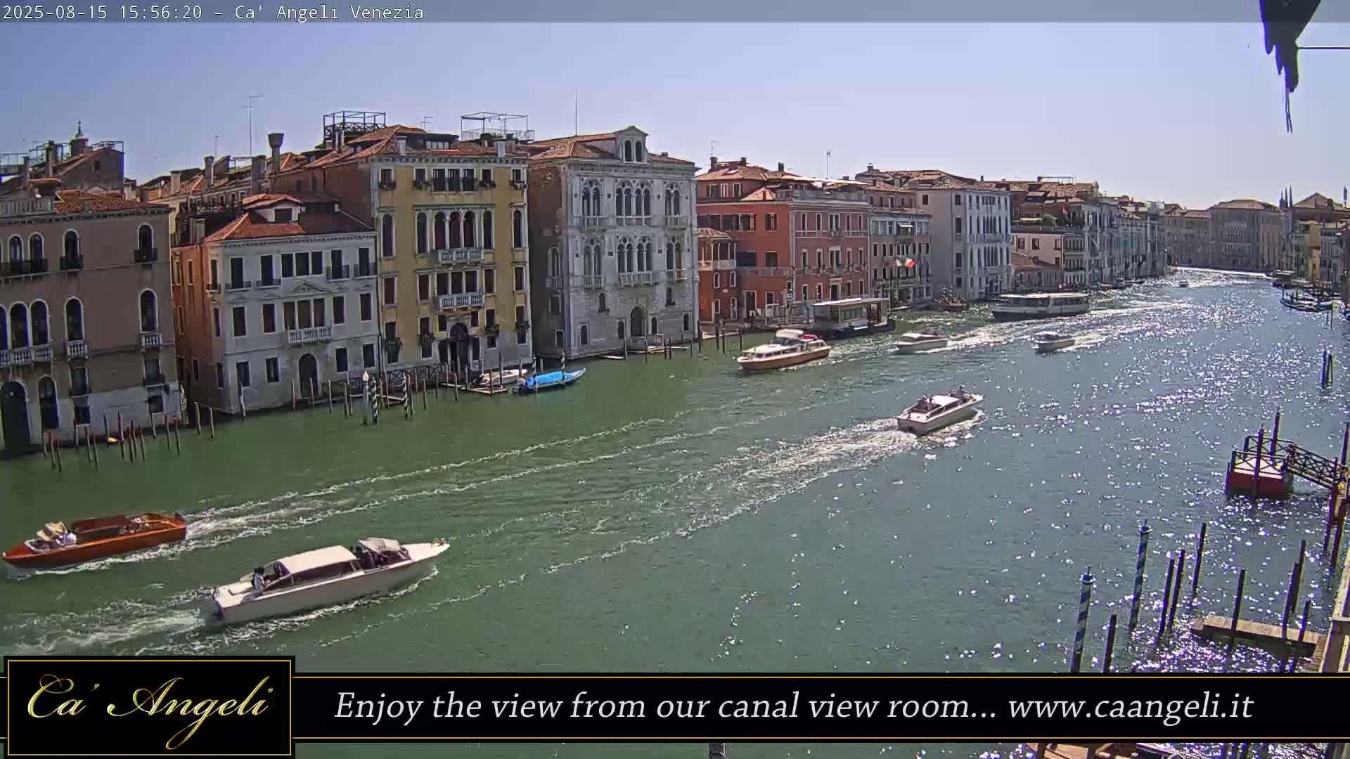 A wide shot of a canal in Venice, Italy, shows several multi-story buildings lining the waterway under a cloudy sky, with various boats moored along the buildings and others traveling on the water.