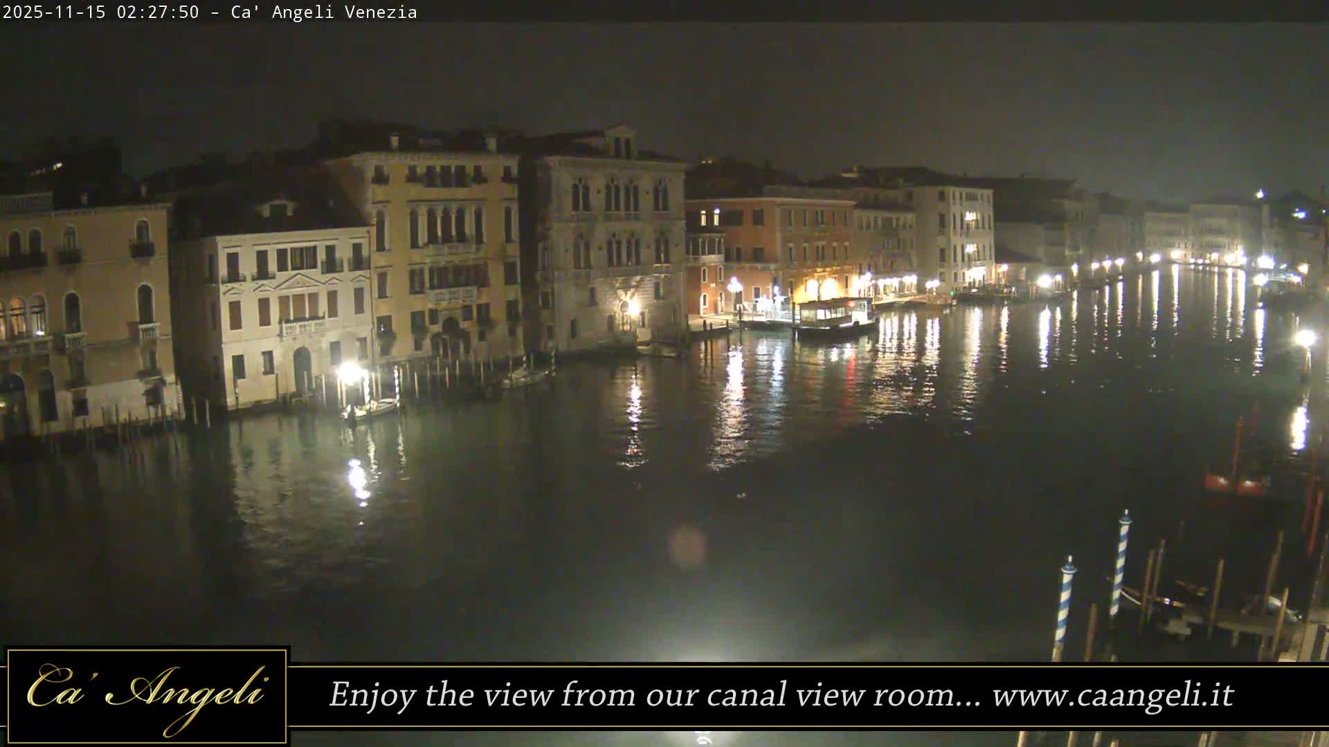 A clear night view of a wide canal lined with illuminated ornate buildings, their bright lights creating shimmering reflections on the calm water, with several boats docked along the edges.