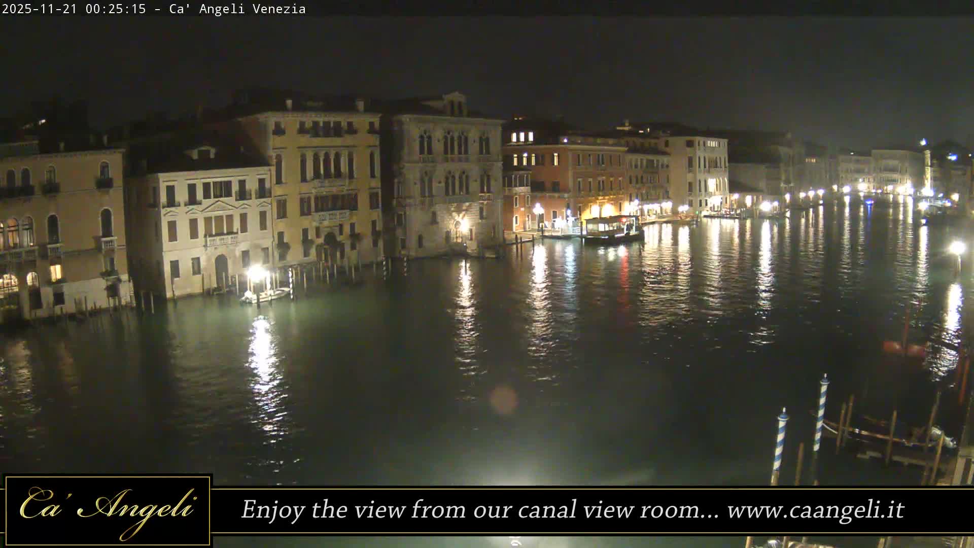 A clear night view of a wide canal lined with illuminated ornate buildings, their bright lights creating shimmering reflections on the calm water, with several boats docked along the edges.