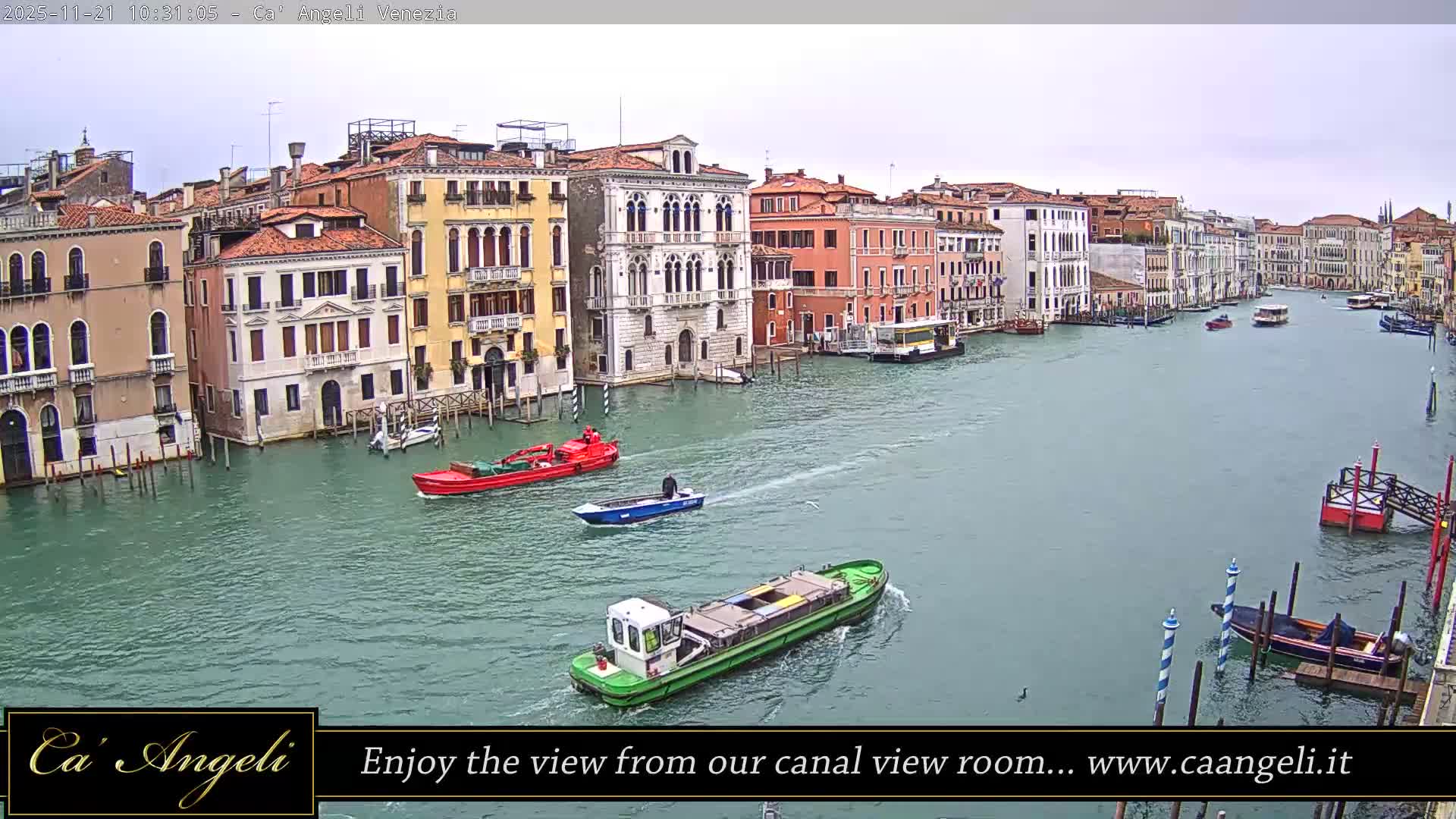 On a cloudy day, the Grand Canal in Venice is bustling with various boats, including barges and smaller vessels, navigating its greenish-blue waters flanked by ornate, colorful historic buildings.