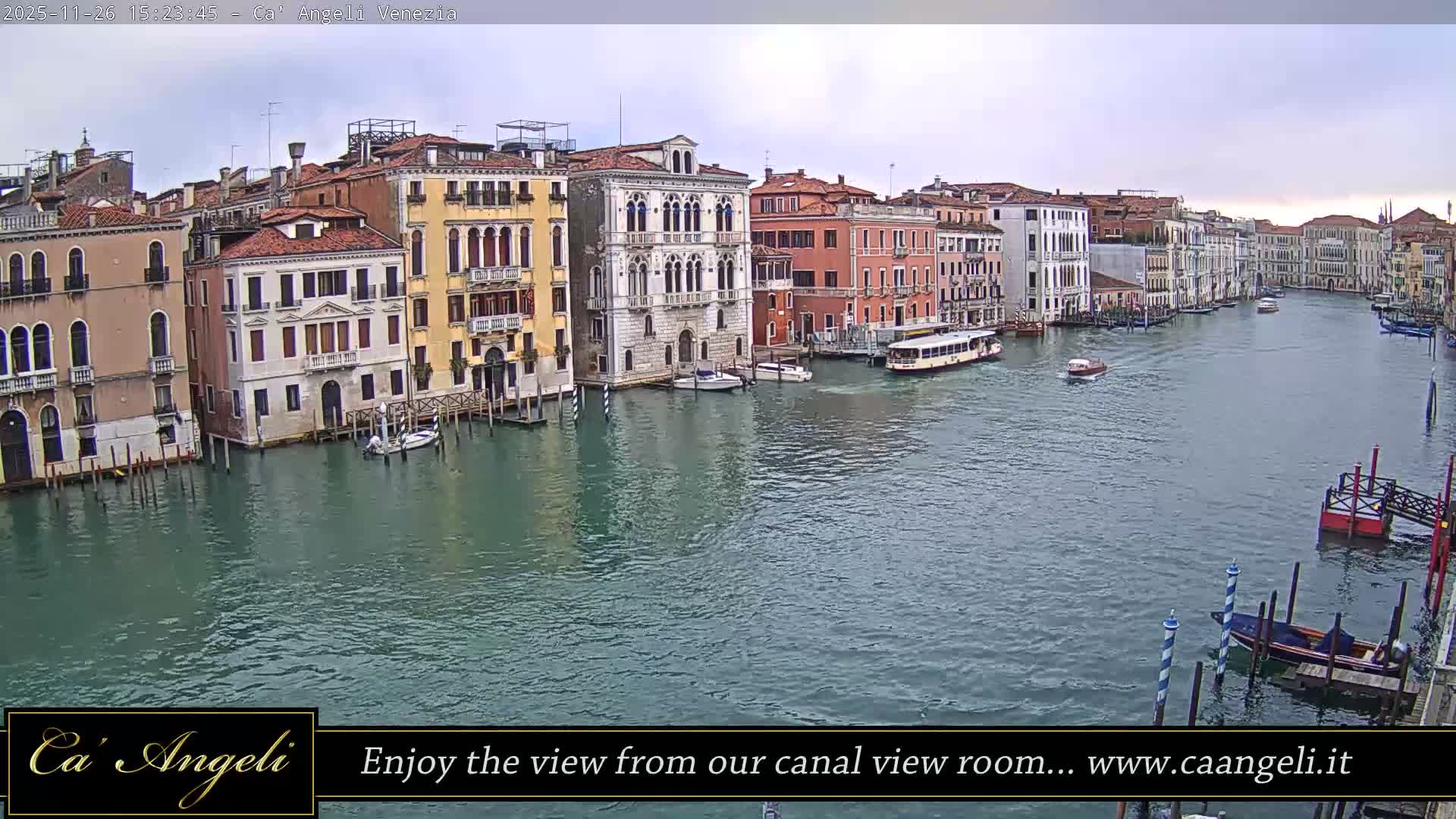 On a cloudy day, the Grand Canal in Venice is bustling with various boats, including barges and smaller vessels, navigating its greenish-blue waters flanked by ornate, colorful historic buildings.