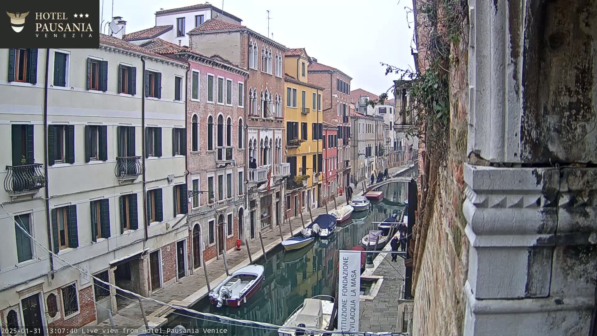 A view down a narrow canal in Venice on an overcast day, showing colorful buildings lining the waterway with several boats moored along the sides.
