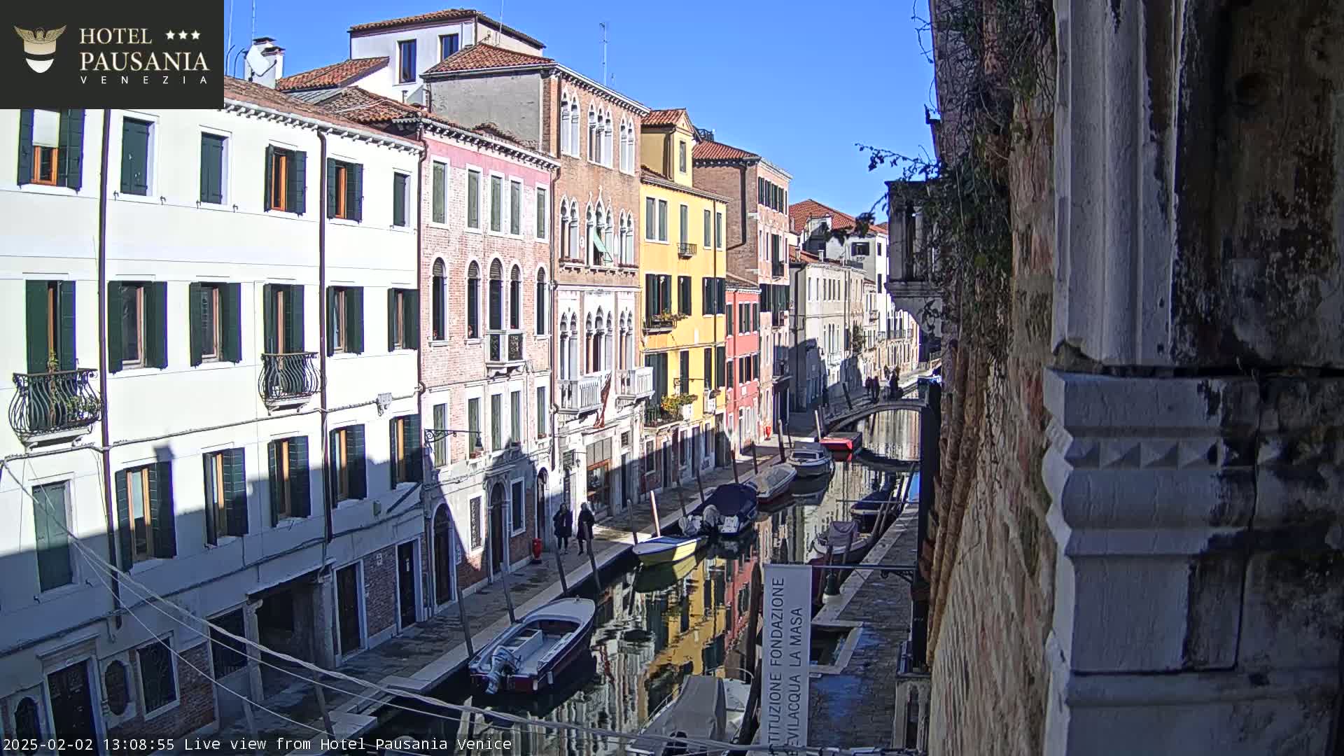 A sunny day in Venice shows a narrow canal lined with colorful buildings, several boats moored along its edge, and pedestrians walking along the canal's bank.