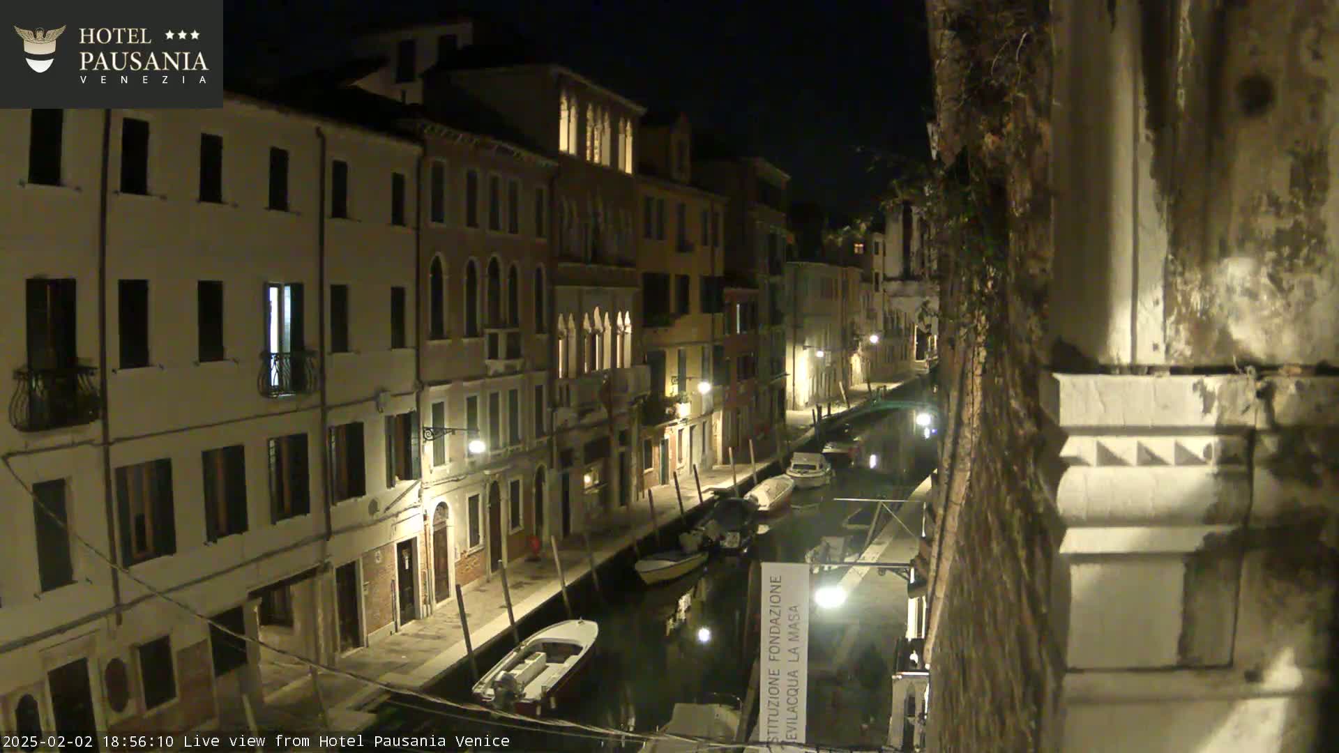 A nighttime view of a narrow canal in Venice, lined with illuminated buildings, with several small boats moored along its edges.