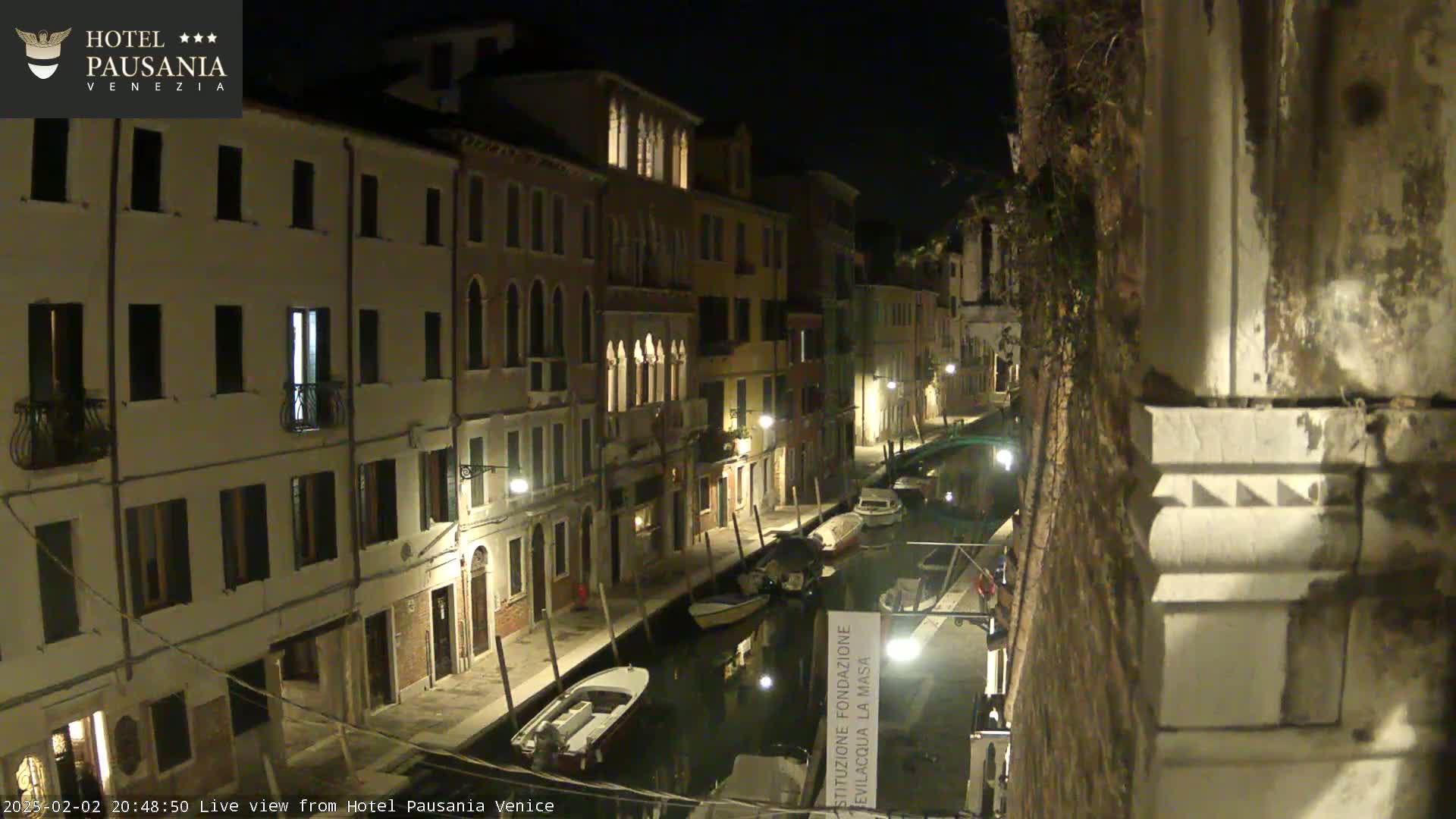 A nighttime view of a narrow canal in Venice, lined with illuminated buildings, with several small boats moored along its banks.