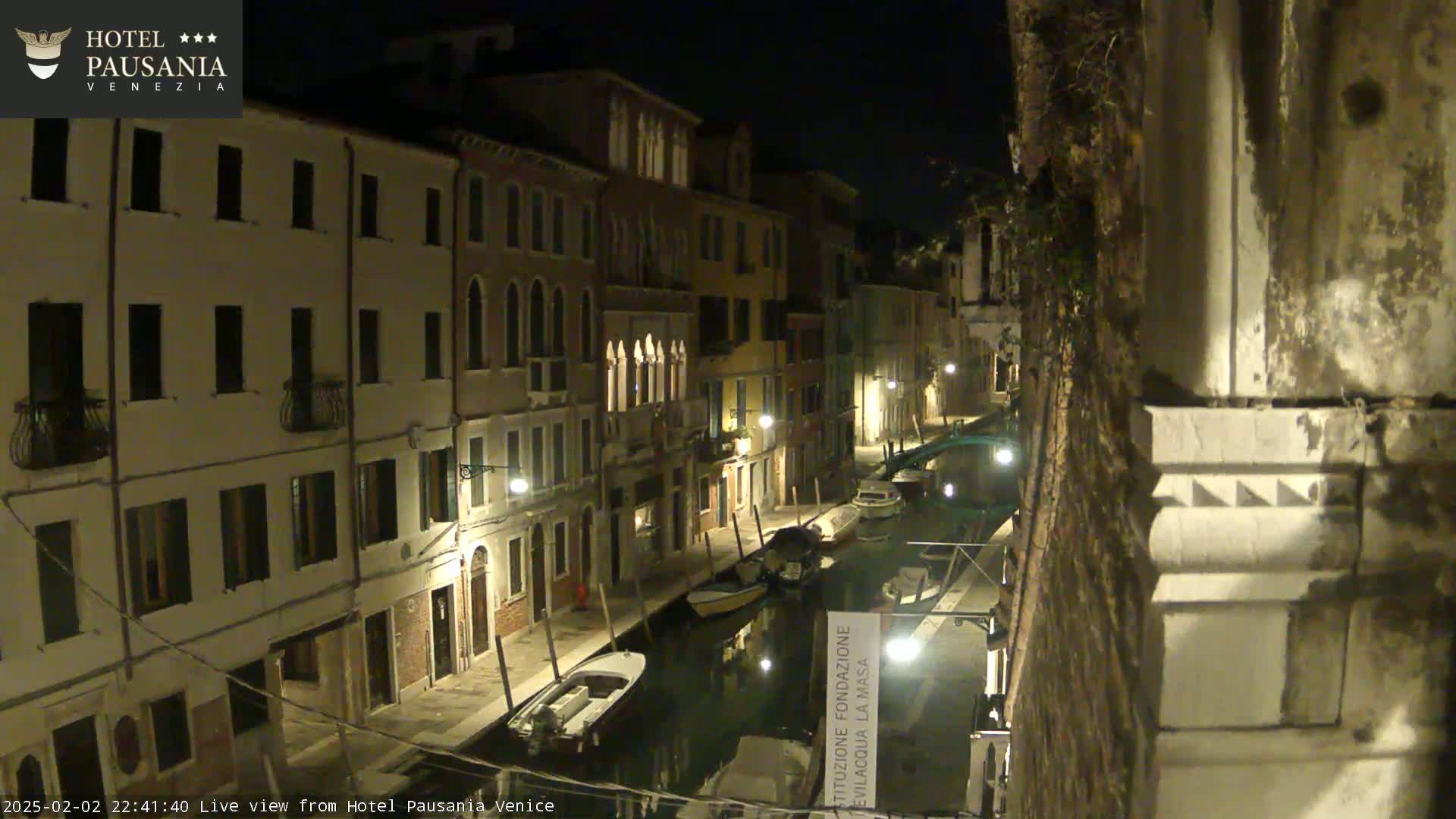 A nighttime view of a narrow canal in Venice, lined with buildings and illuminated by streetlights, with several boats moored along its banks.