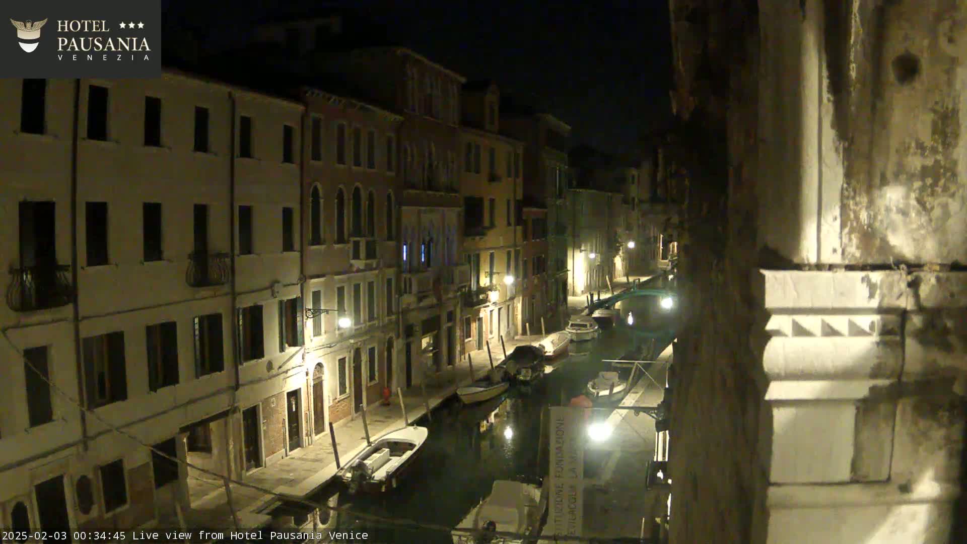 At night, several boats are moored along a canal lined with illuminated buildings.