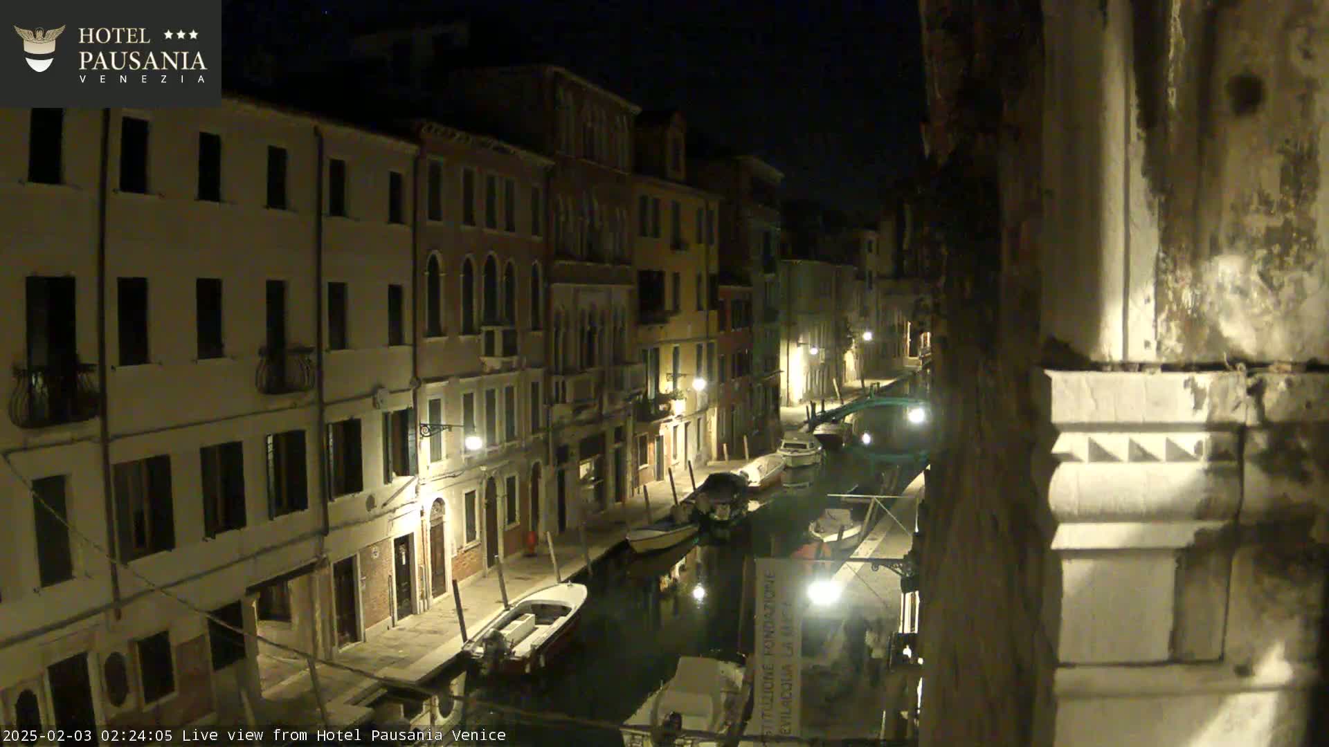 A nighttime view of a narrow canal in Venice, lined with buildings illuminated by streetlights, showing several moored boats.