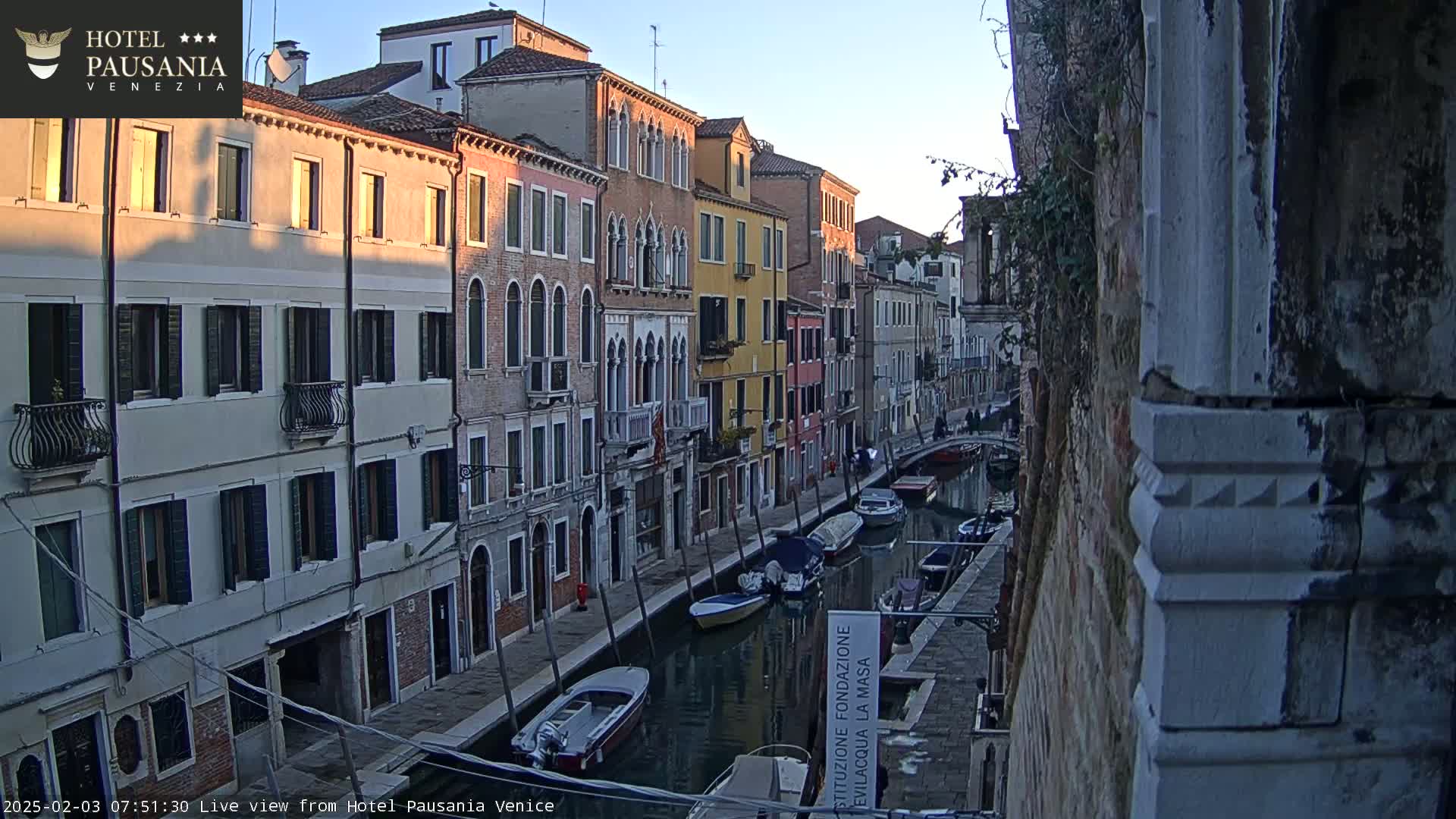 A narrow Venetian canal at sunrise is lined with closely-packed buildings, several small boats are moored along its sides.