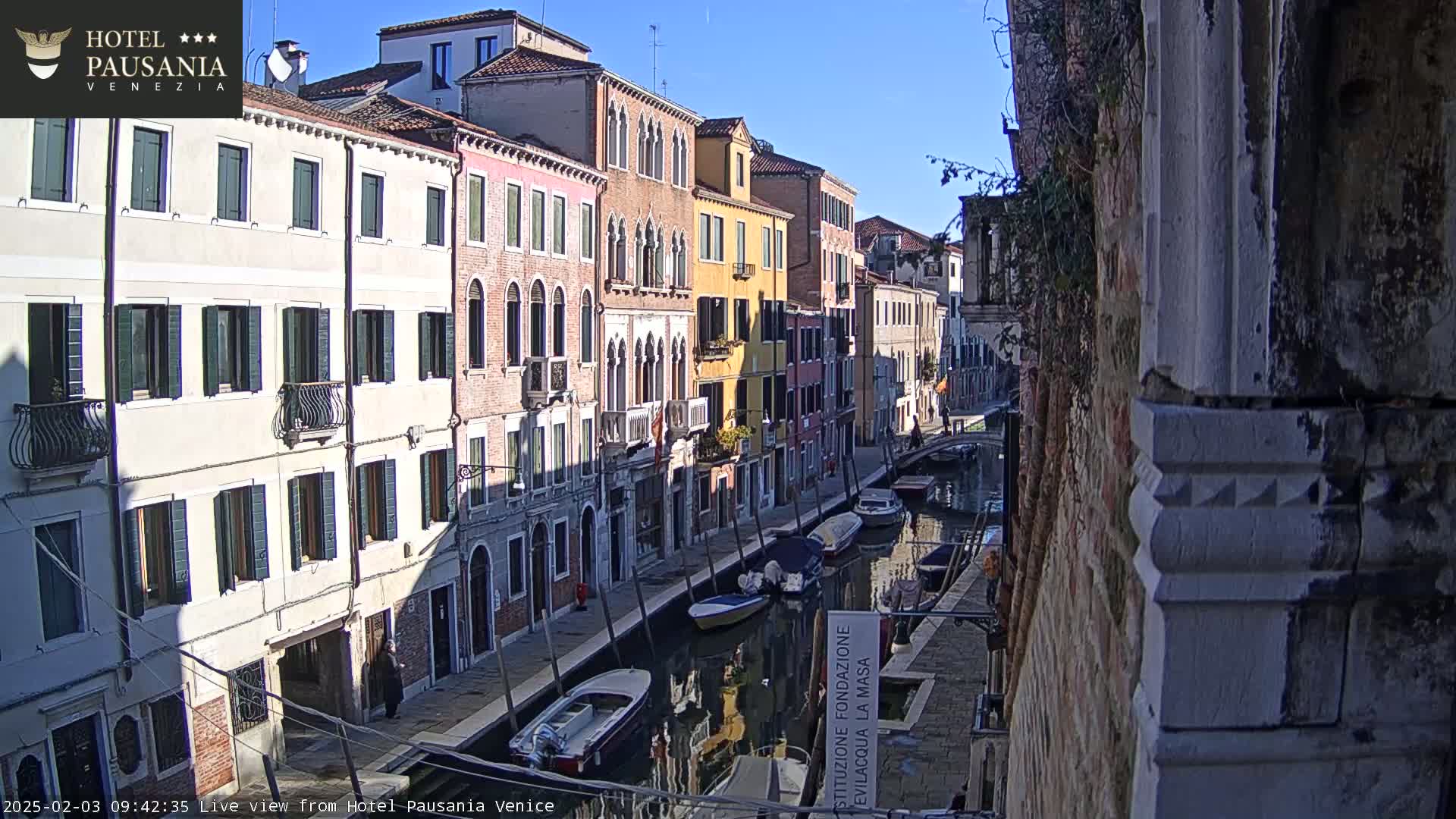 A sunny day in Venice shows several colorful buildings lining a canal with boats moored along the waterway.