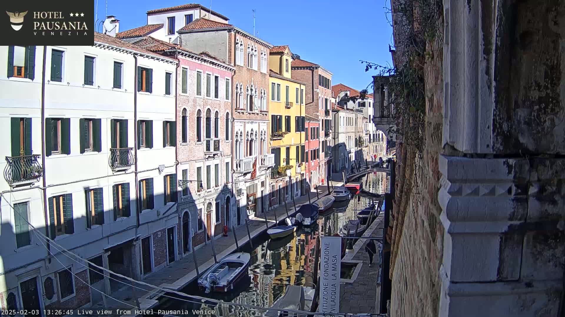 A sunny day in Venice shows a narrow canal lined with colorful buildings and several moored boats.
