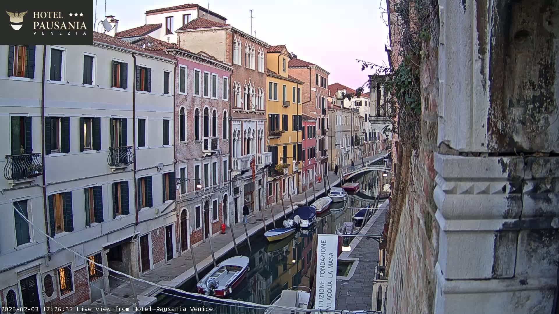 A narrow canal in Venice, lined with colorful buildings, is seen at dusk with several boats moored along its sides.