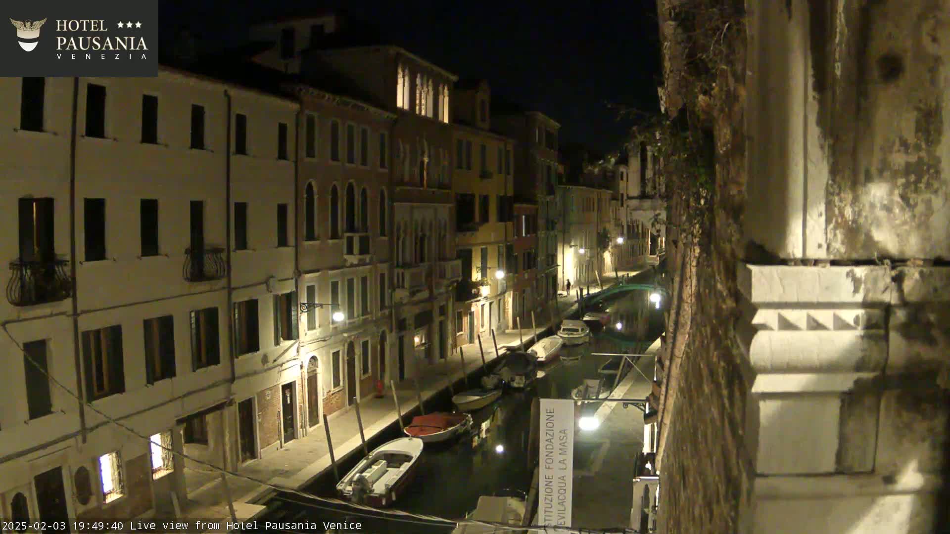 A nighttime view of a narrow canal in Venice, lined with illuminated buildings and several moored boats.