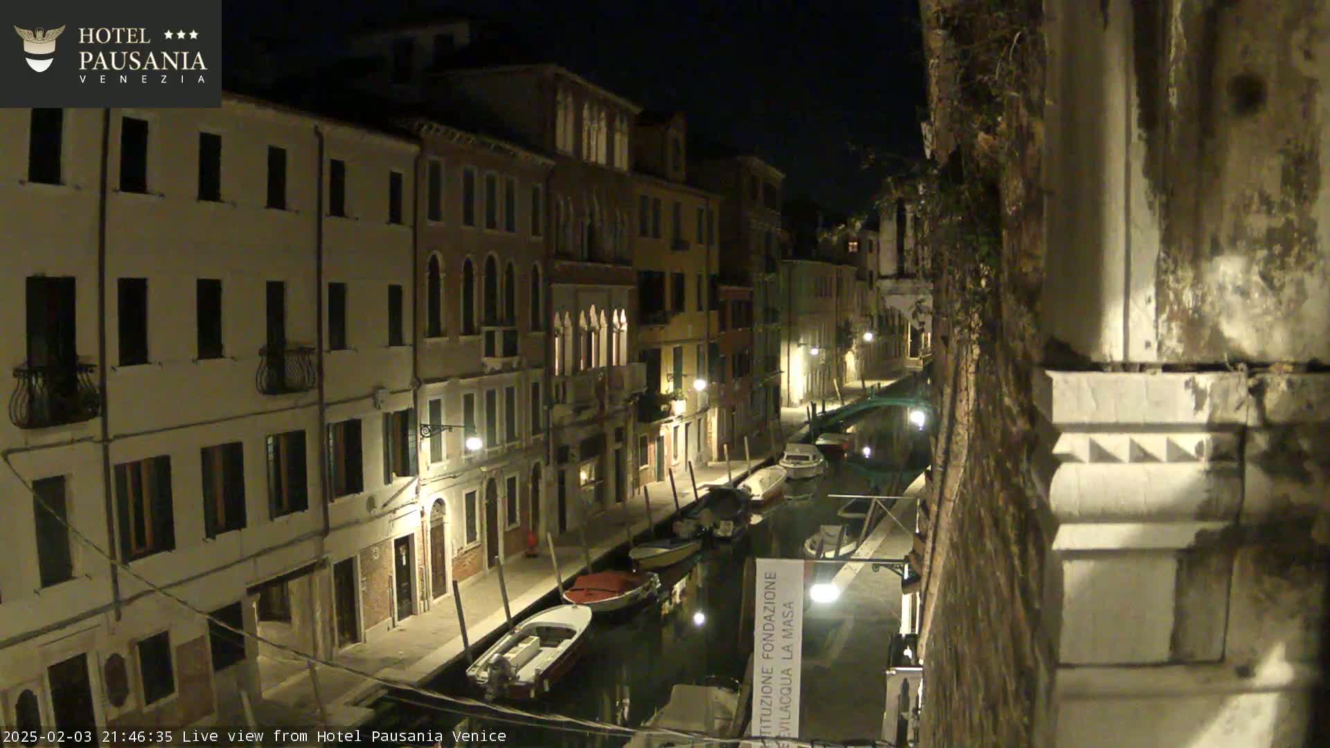 A nighttime view of a narrow canal in Venice, lined with illuminated buildings and several moored boats.