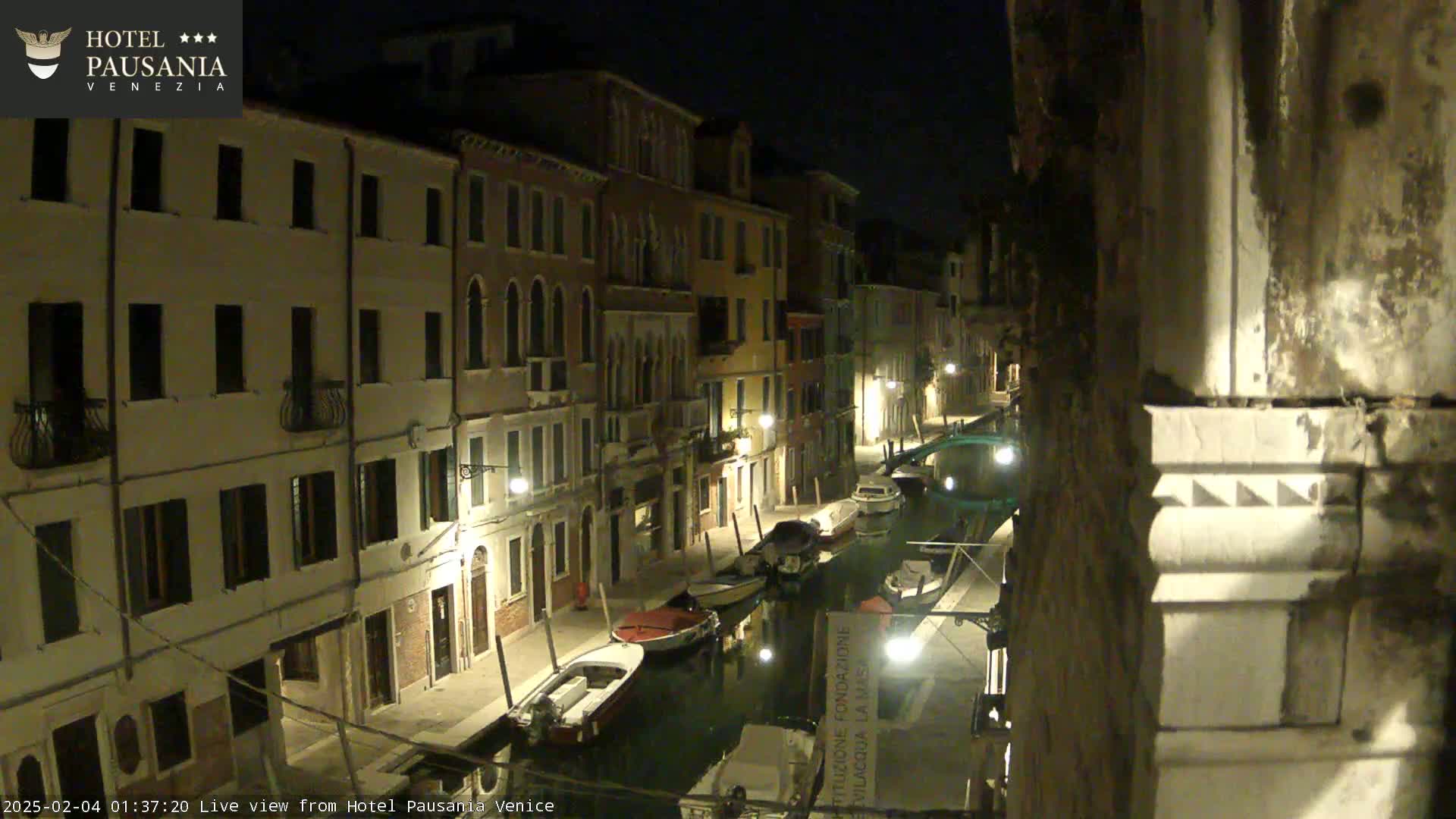A nighttime view of a narrow canal in Venice, lined with illuminated buildings and several moored boats.