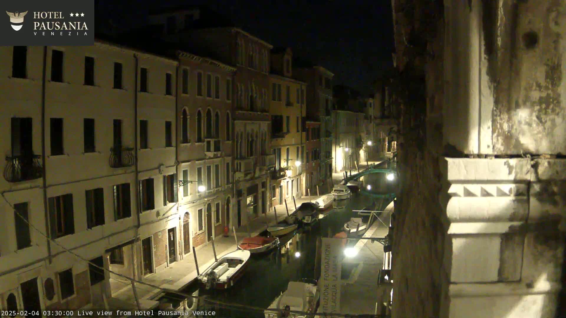 A nighttime view of a narrow canal in Venice, lined with illuminated buildings and several moored boats.
