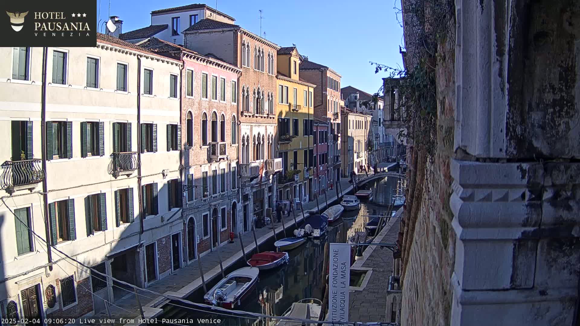 A sunny day in Venice shows a narrow canal lined with colorful buildings and several small boats moored along the waterway.