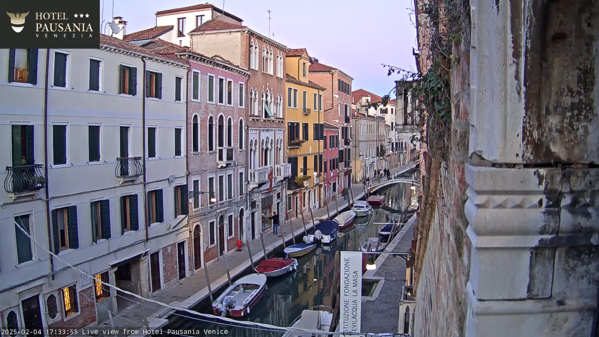 A narrow canal in Venice, Italy, lined with colorful buildings, shows several boats moored along its sides under a twilight sky.