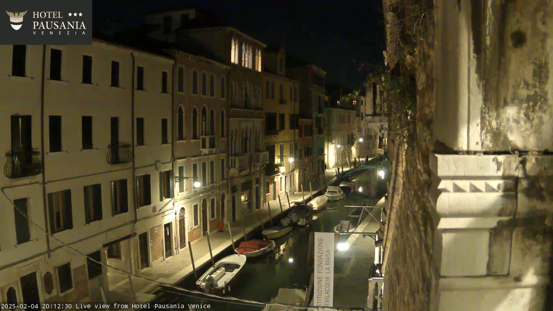 A nighttime view of a narrow canal in Venice, lined with illuminated buildings and several moored boats.