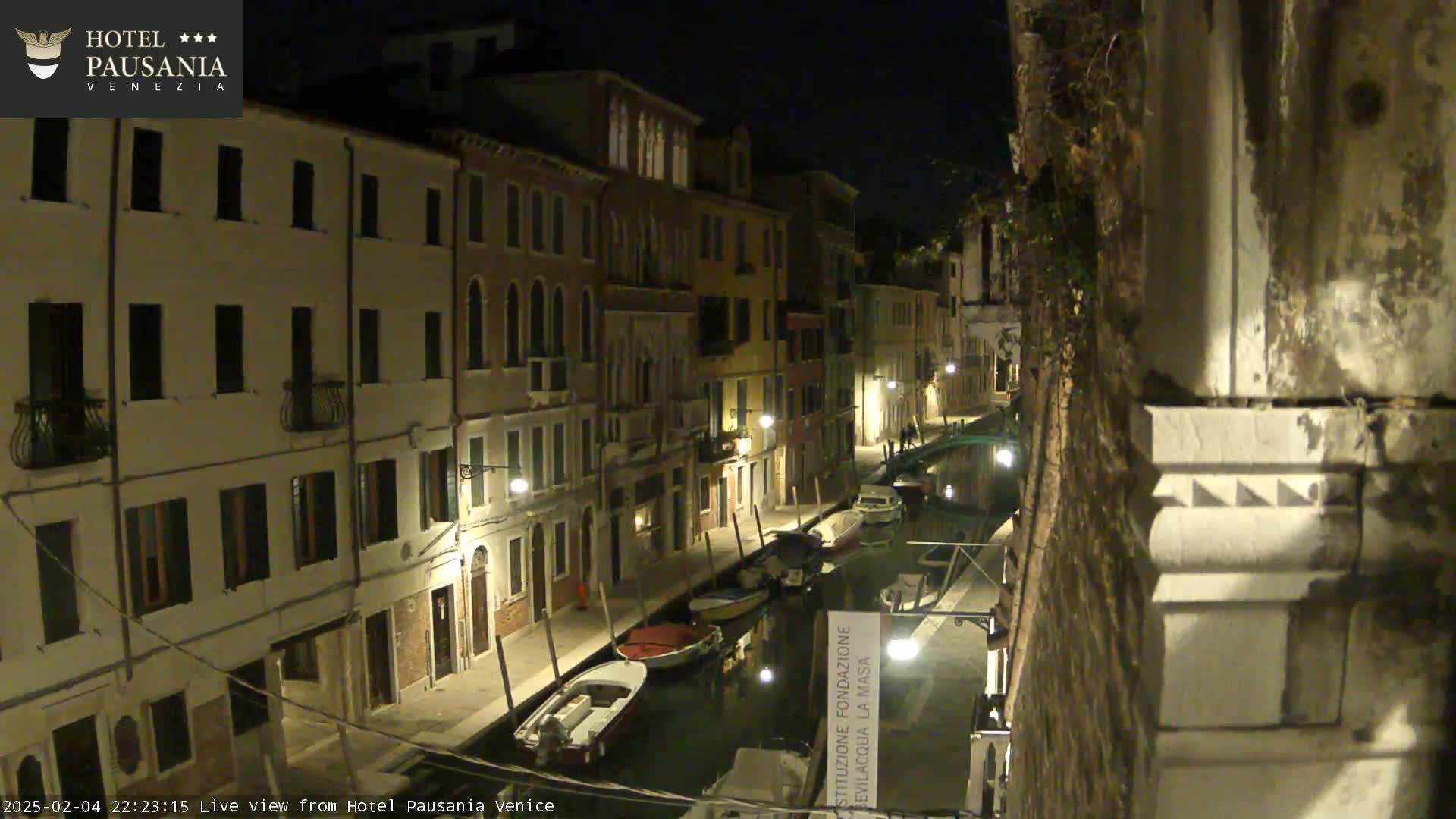 A nighttime view of a narrow Venetian canal lined with buildings, showing several boats moored along the waterway under dark, clear skies.