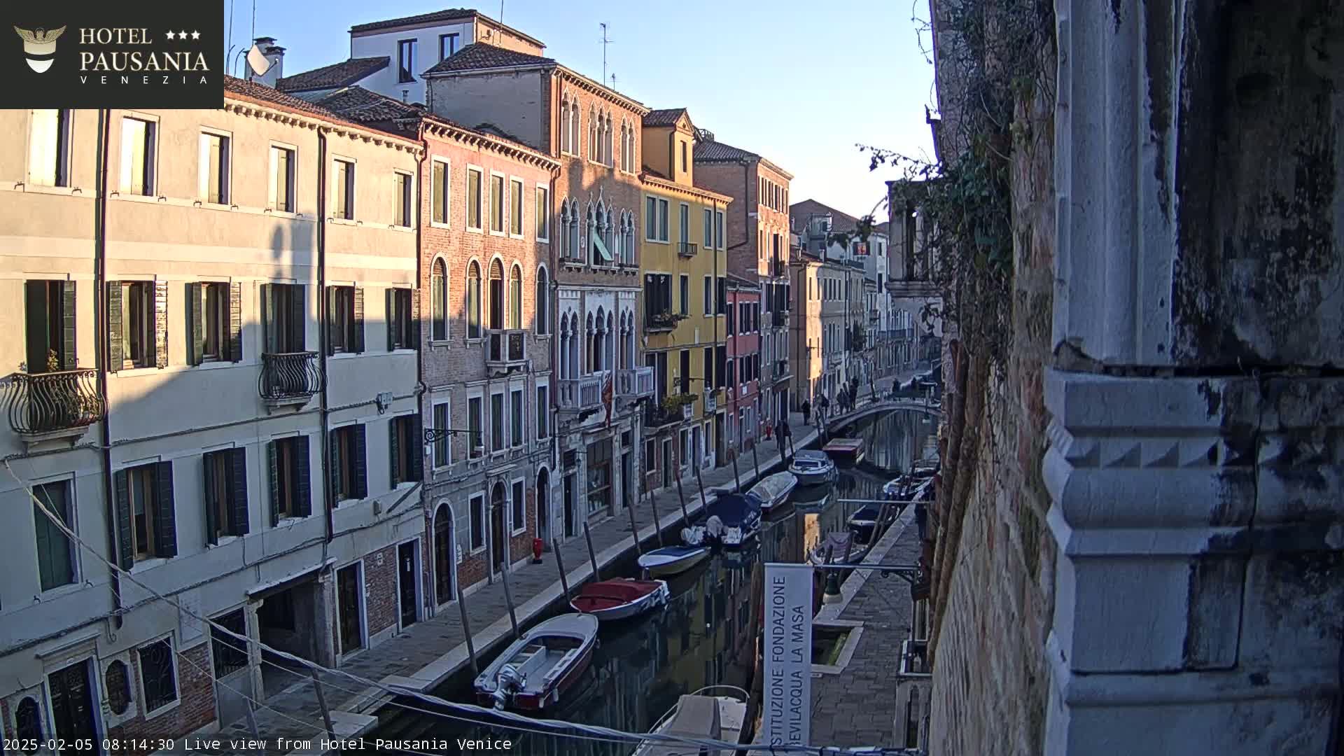 A narrow Venetian canal, lined with colorful buildings under a clear, bright sky, features several moored boats.