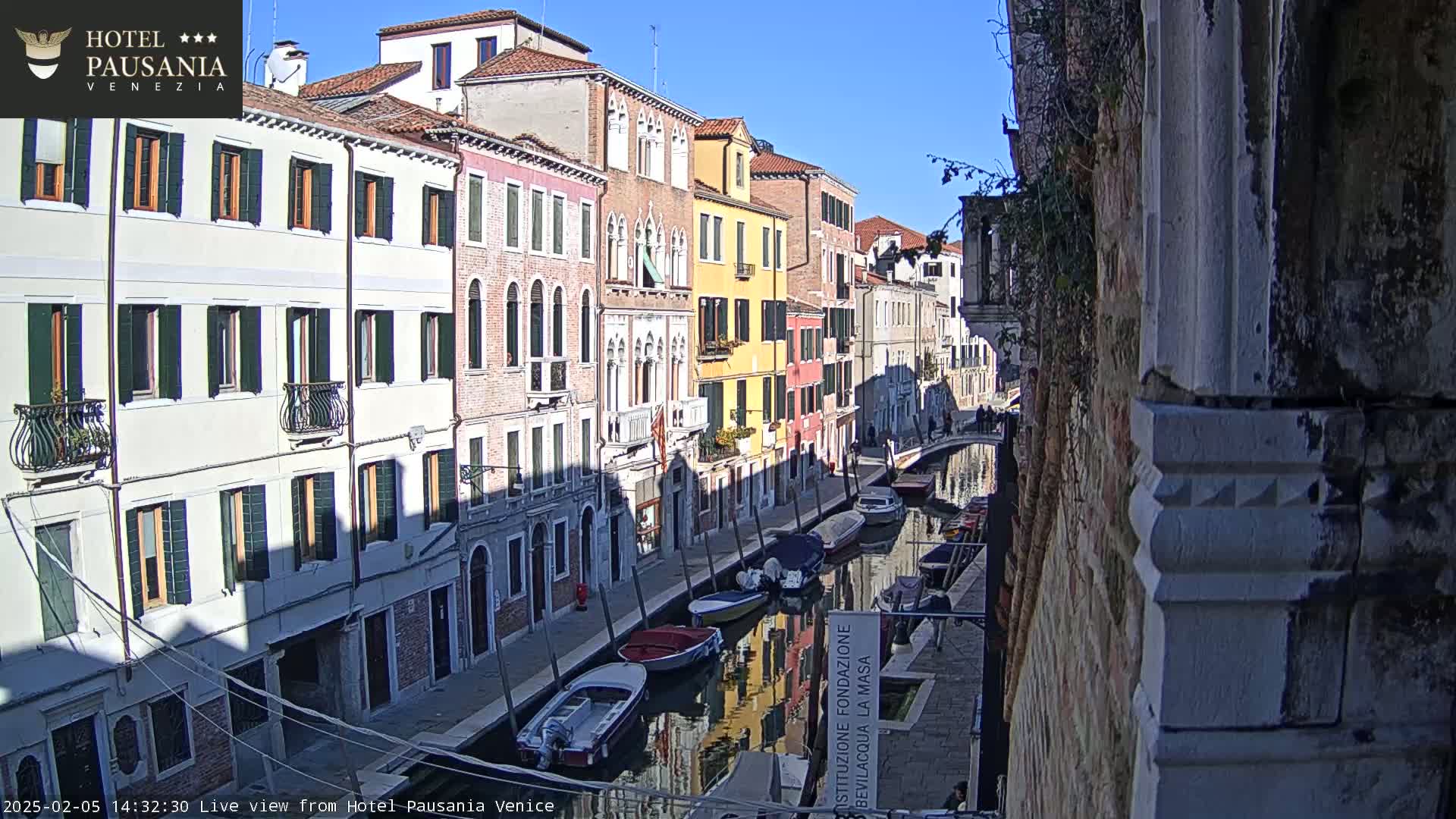 A sunny day in Venice shows a narrow canal lined with colorful buildings, several boats moored along the waterway.