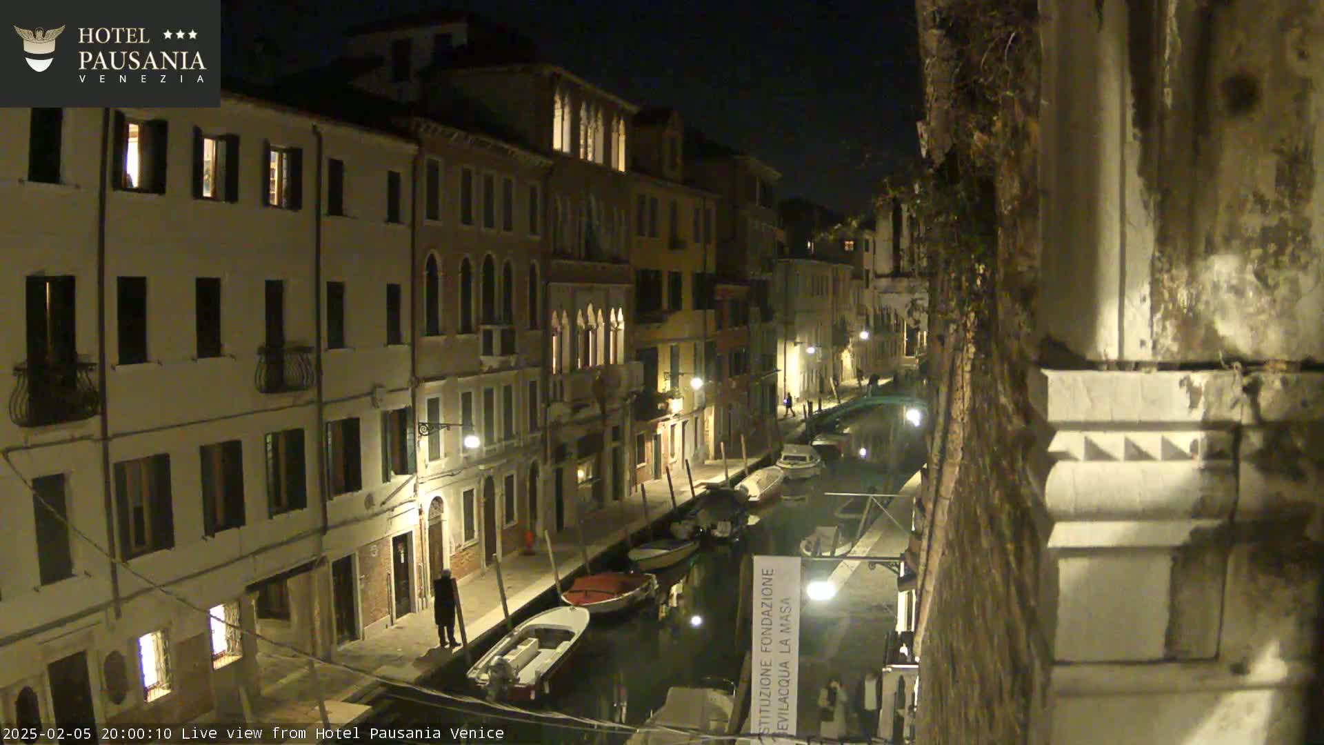 A nighttime view of a canal in Venice, Italy, lined with illuminated buildings and several moored boats.