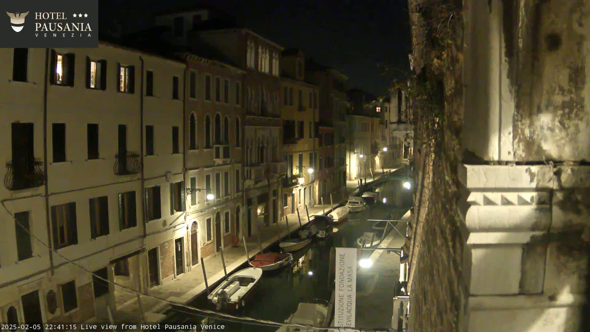 A nighttime view of a narrow canal in Venice, lined with illuminated buildings and several small boats moored along its sides.