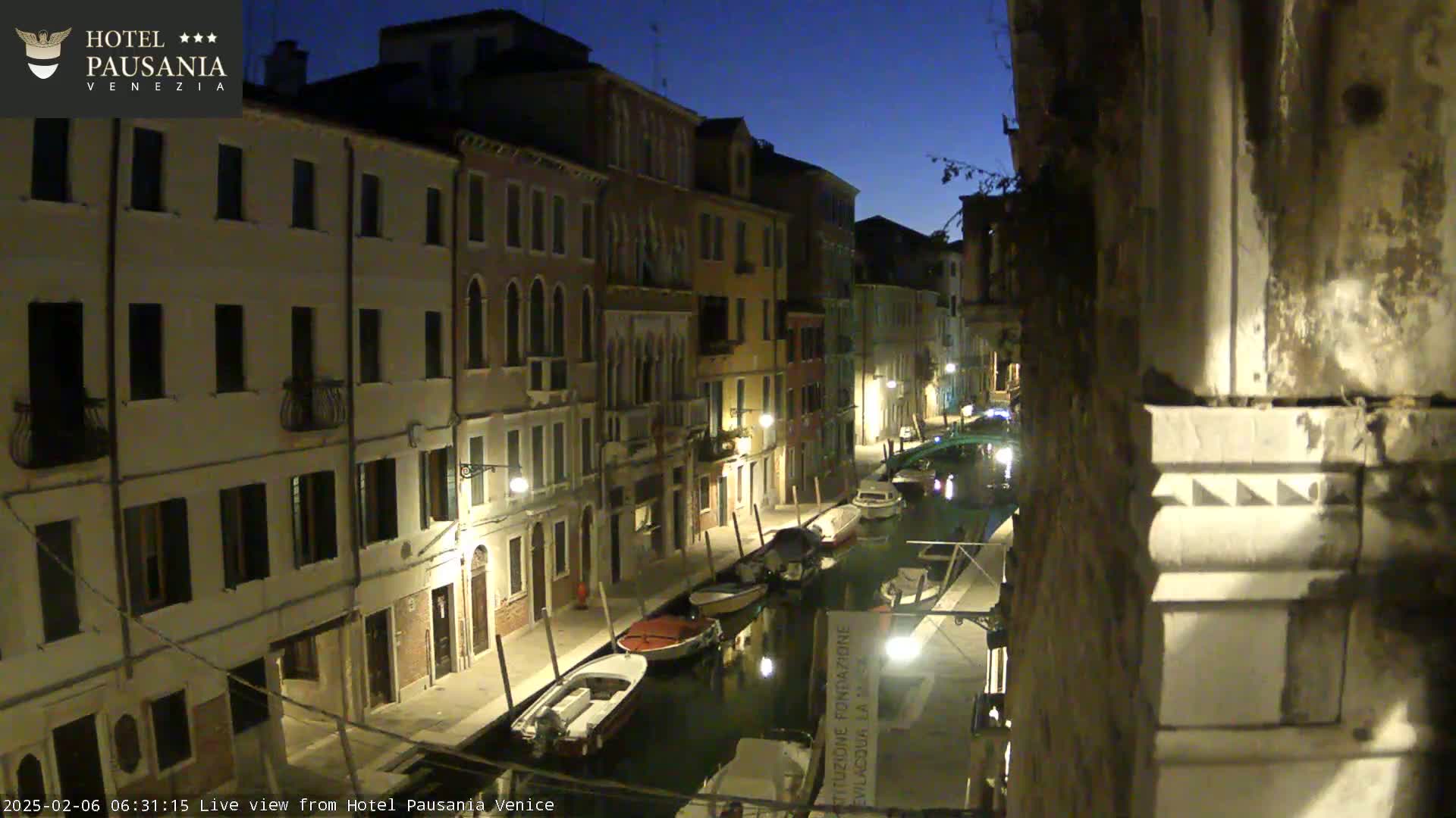 A nighttime view of a narrow canal in Venice, lined with buildings and several moored boats under a dark blue sky.