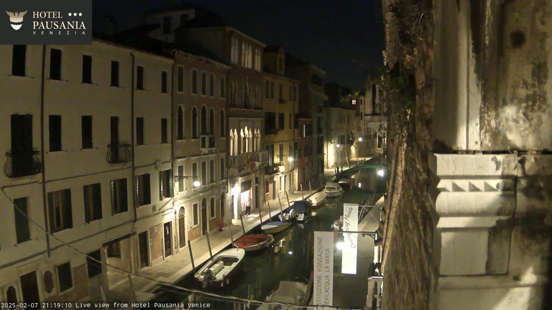 A nighttime view of a narrow canal in Venice, lined with illuminated buildings and several small boats moored along its banks.