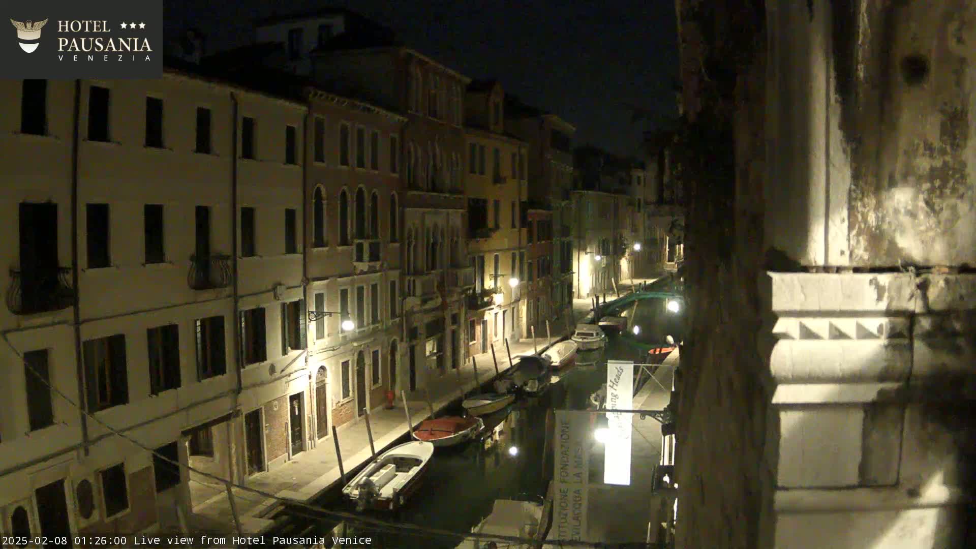 A nighttime view of a narrow canal in Venice, lined with illuminated buildings and several moored boats.