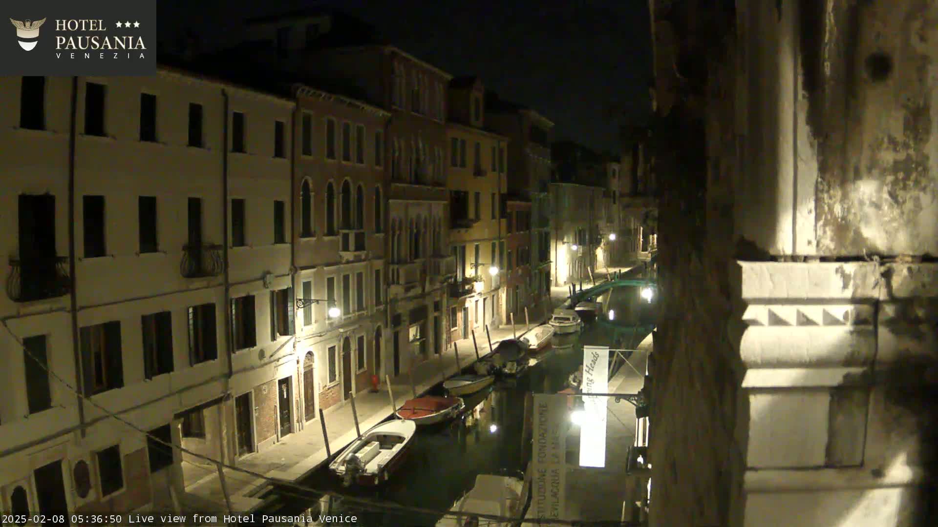 A nighttime view of a narrow canal in Venice, lined with buildings and several moored boats.