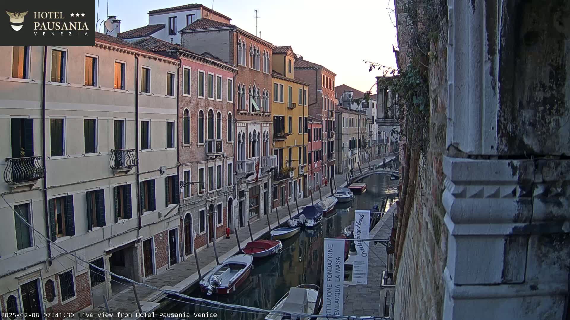 A calm canal in Venice, lined with colorful buildings, is filled with several moored boats under a sunrise sky.