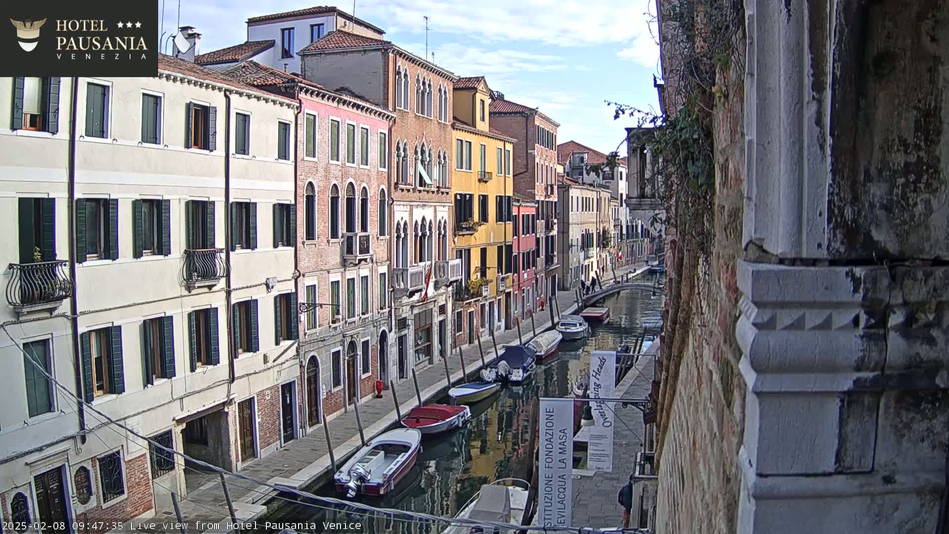 A canal in Venice, under a partly cloudy sky, is lined with colorful buildings and several moored boats.