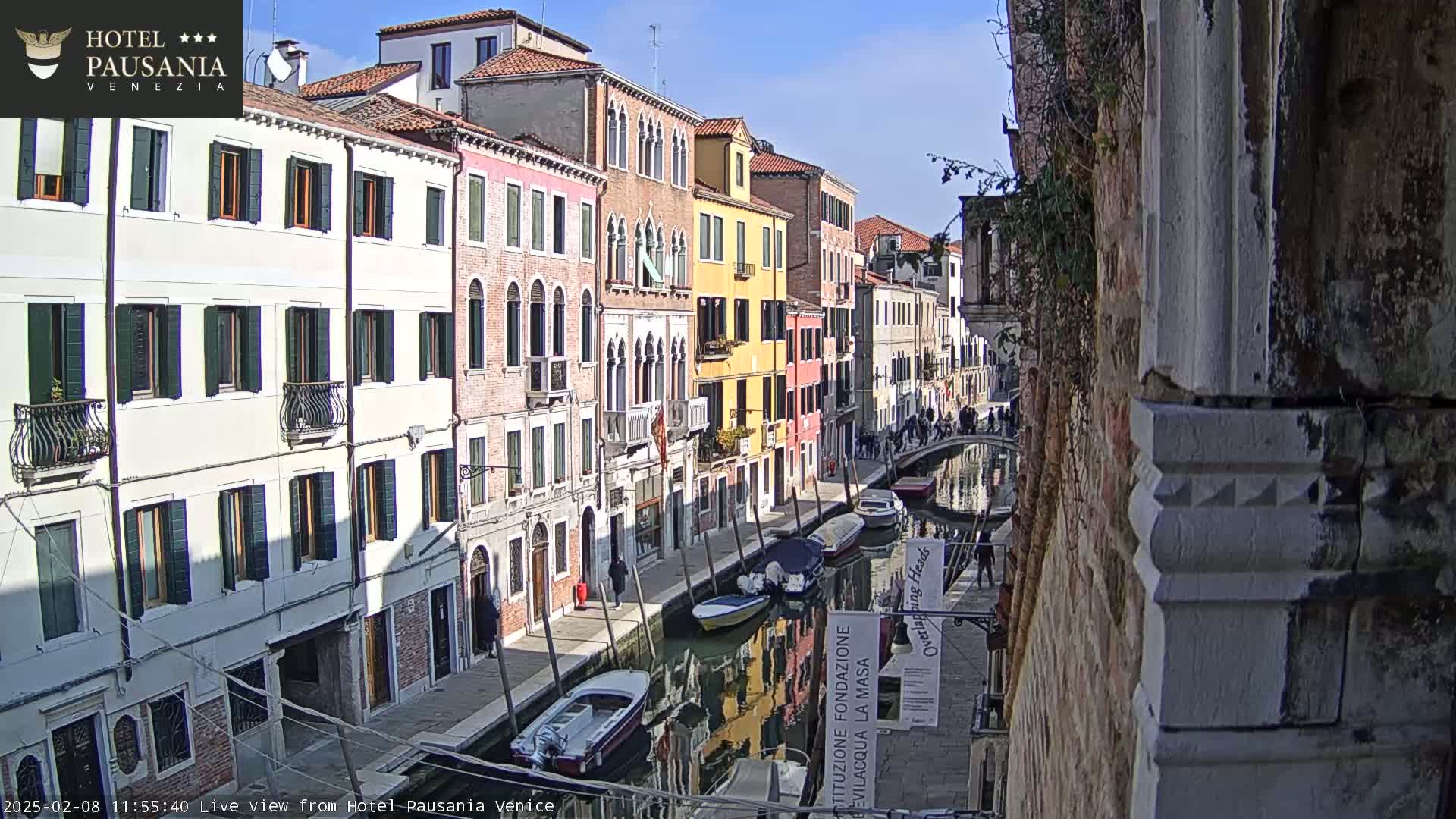 A sunny day reveals a narrow Venetian canal lined with colorful buildings, several small boats are moored along its banks, and a few pedestrians stroll along the waterfront.