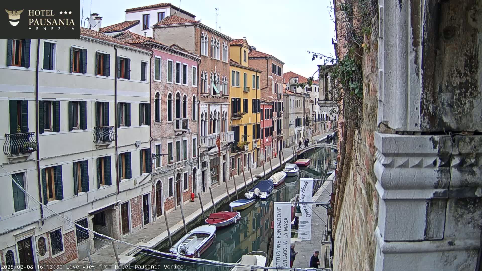 A narrow Venetian canal is lined with colorful buildings under an overcast sky, several boats are moored along the waterway.