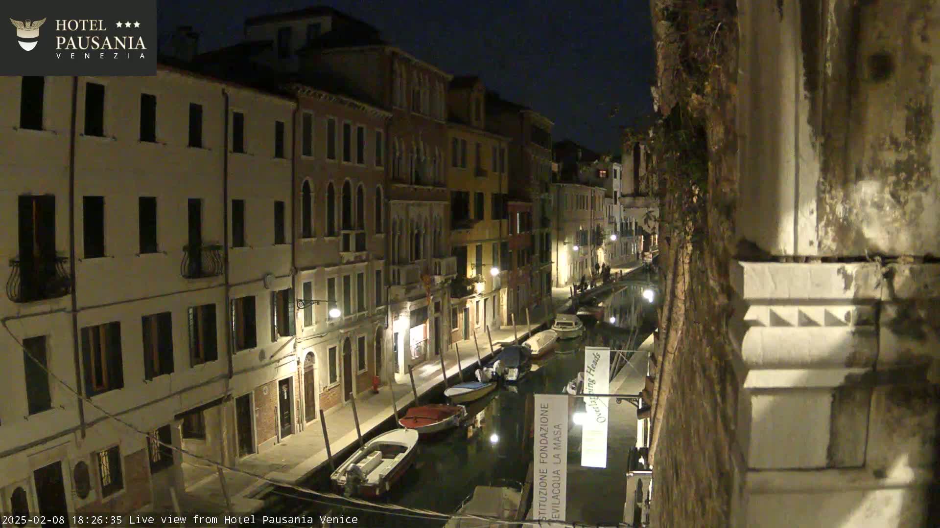 A nighttime view of a canal in Venice, showing several boats moored along the waterway, flanked by illuminated buildings.