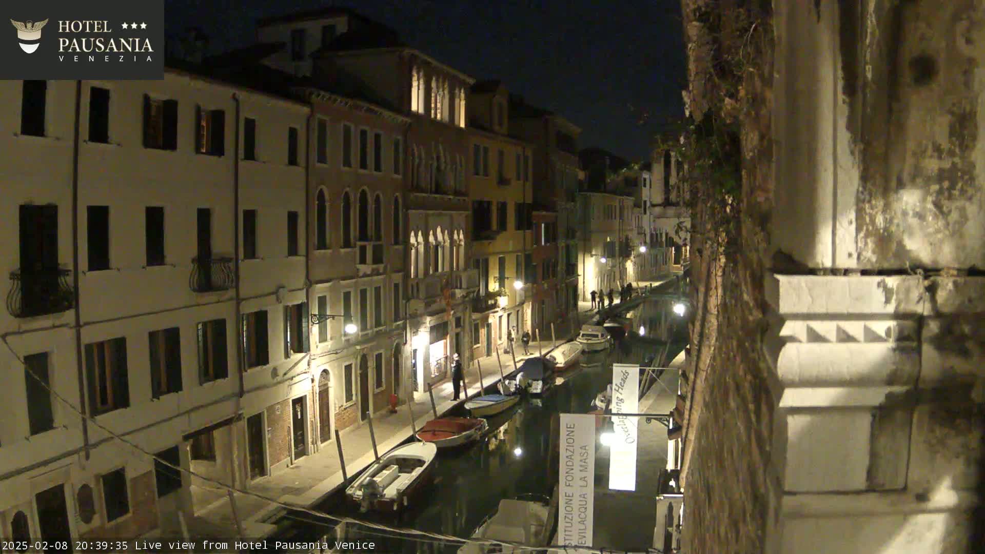 A nighttime view of a narrow canal in Venice, lined with illuminated buildings and several moored boats.