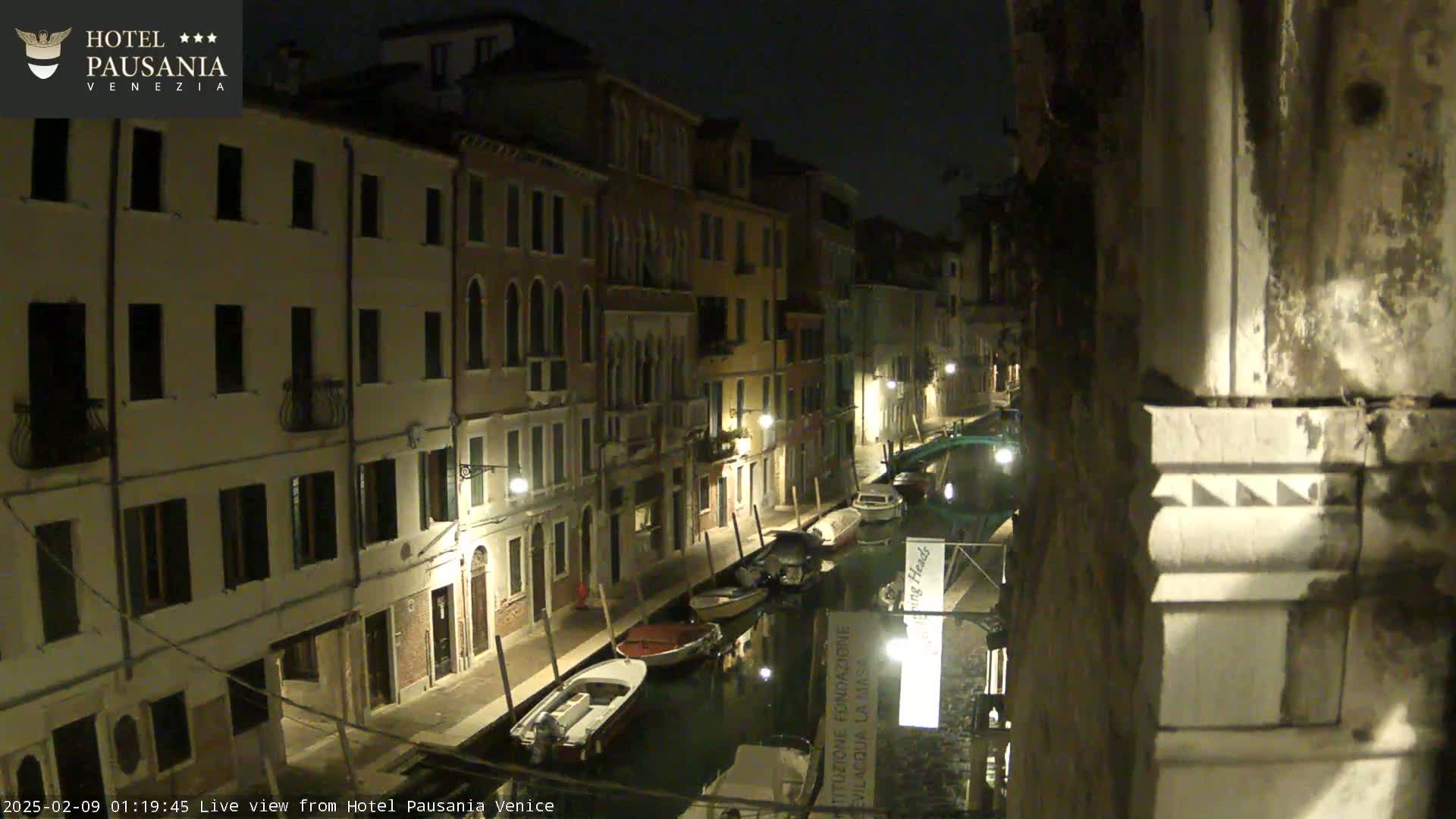 A nighttime view of a narrow canal in Venice, lined with buildings and moored boats, under a dark sky.
