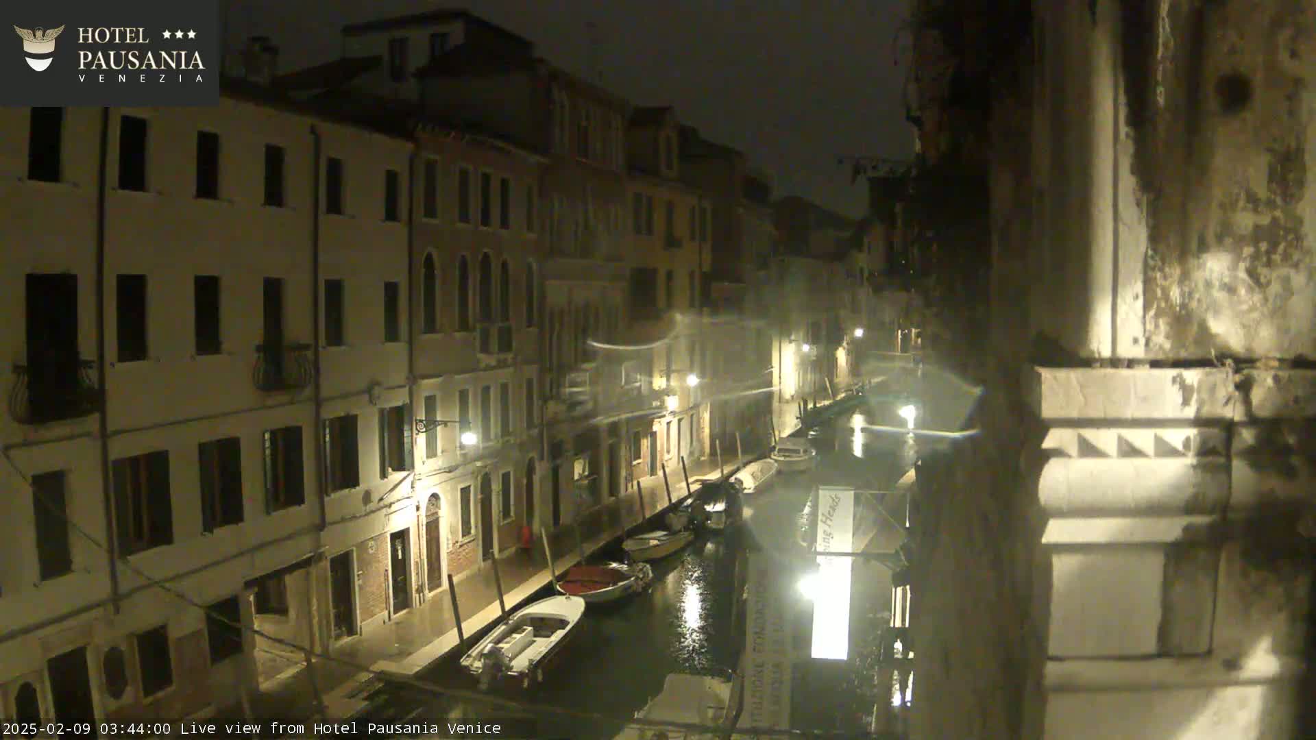 A nighttime view of a canal in Venice, with several boats moored along its banks, flanked by illuminated buildings.