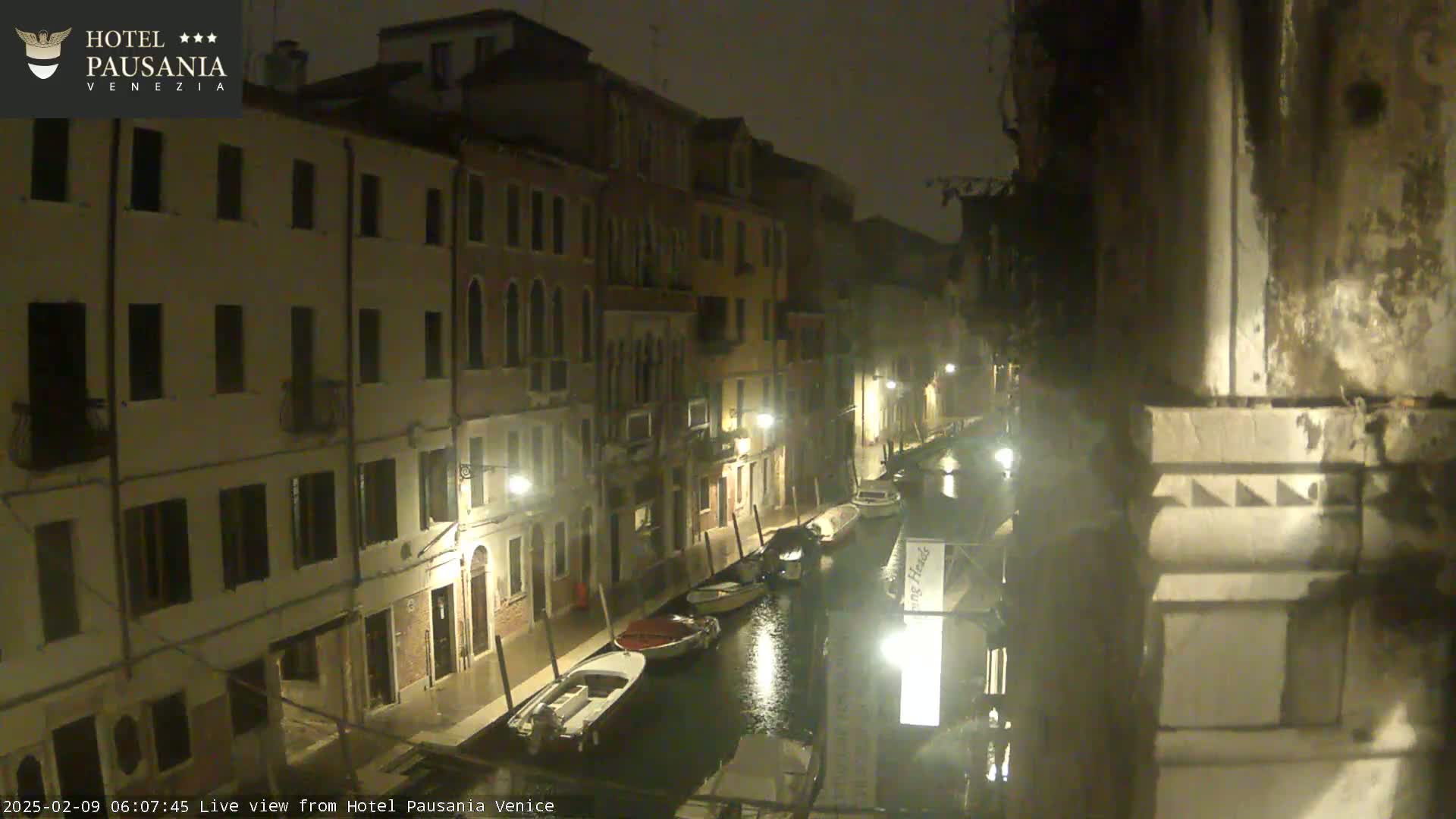 A nighttime view of a narrow canal in Venice, Italy, showing several boats moored along the sides and buildings lining the waterway under a light rain.
