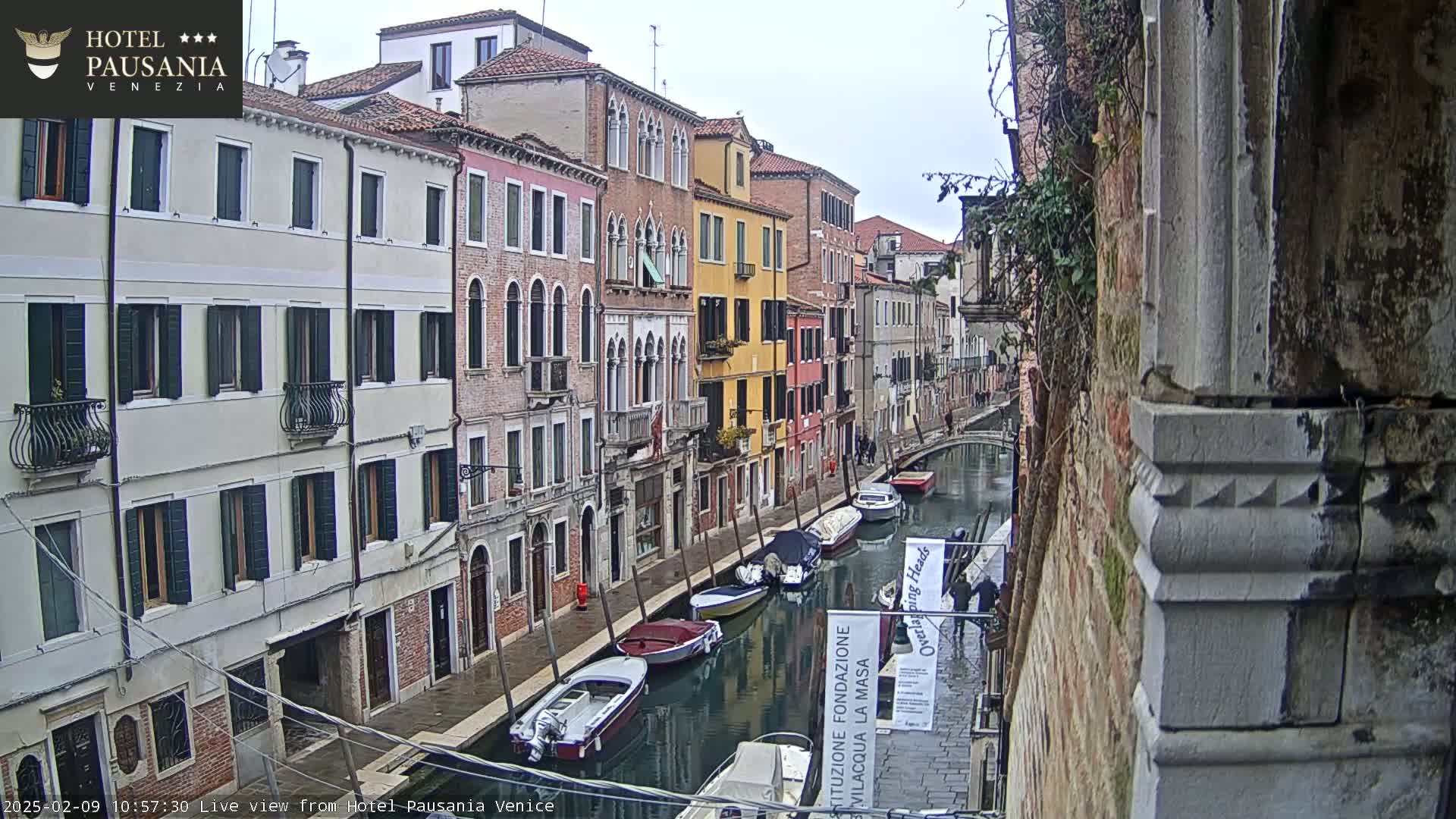 A narrow canal in Venice, Italy, is lined with tall, colorful buildings under an overcast sky, and several boats are moored along its sides.
