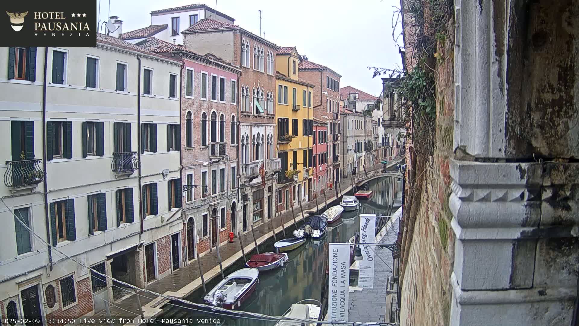 A view of a narrow canal in Venice on an overcast day, lined with closely-spaced multi-story buildings, several small boats are moored along the waterway.