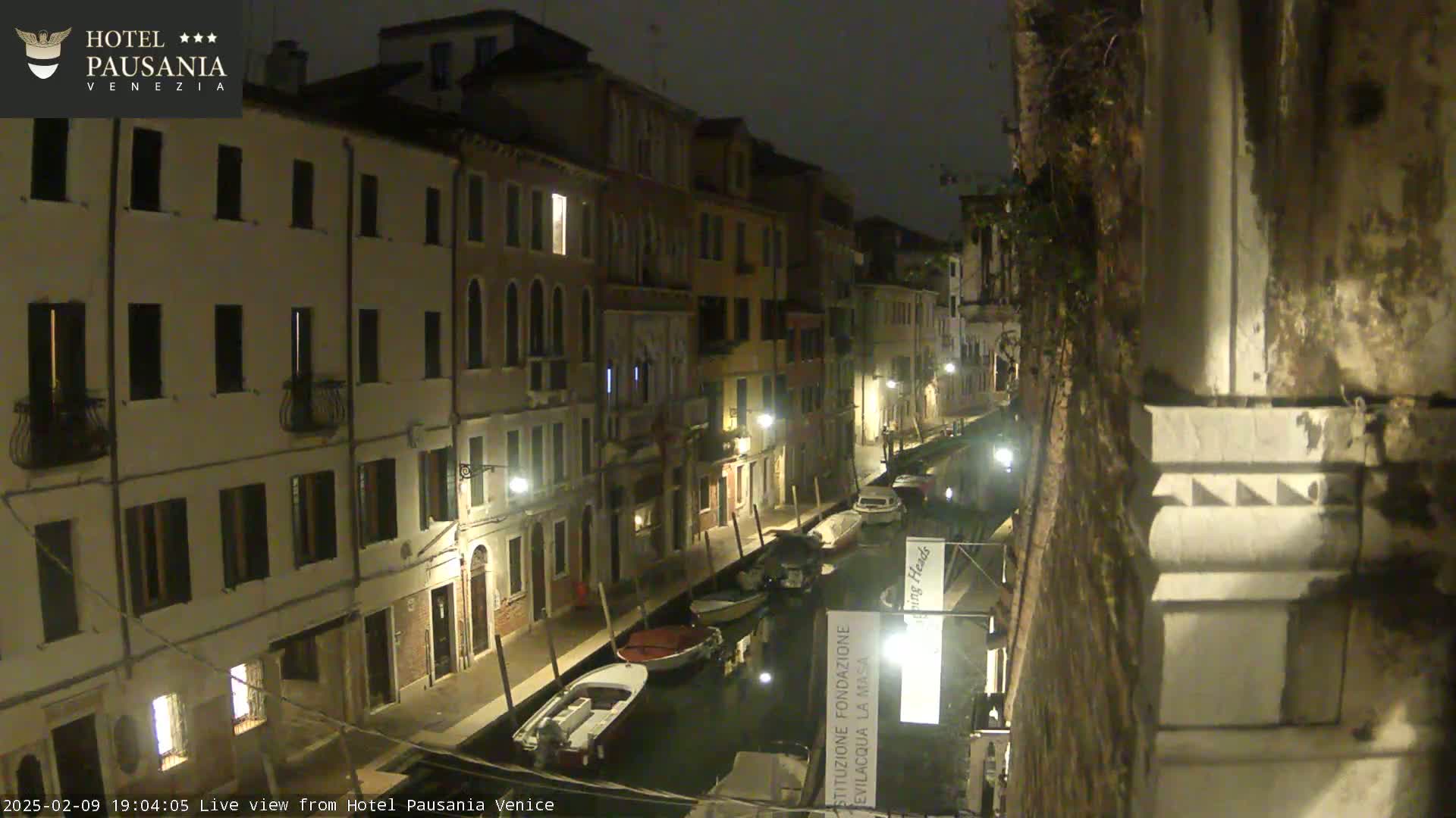 A nighttime view of a narrow canal in Venice, lined with buildings and illuminated by streetlights, showing several boats moored along its banks.