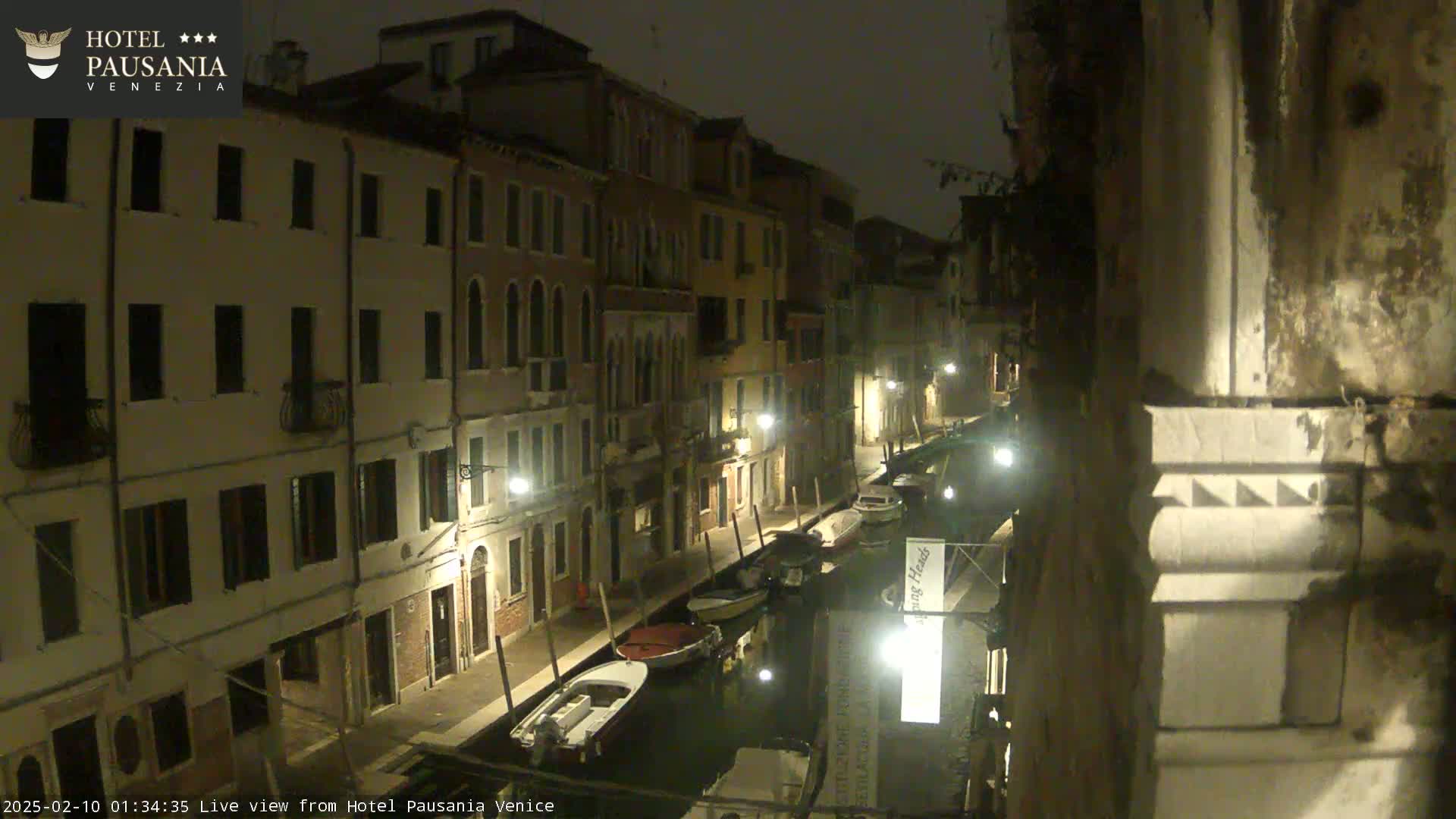 A nighttime view of a narrow canal in Venice, with several boats moored alongside buildings lit by streetlights under a dark, possibly overcast sky.