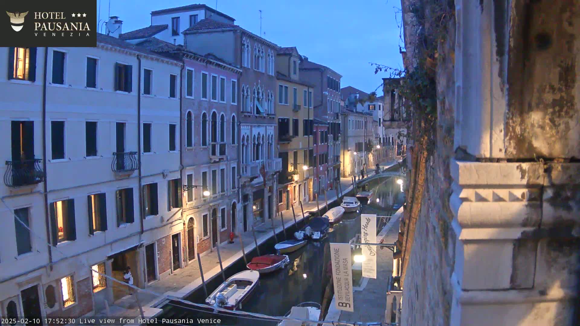 A twilight view of a narrow canal in Venice, Italy, lined with multi-story buildings, and several boats are moored along its banks.